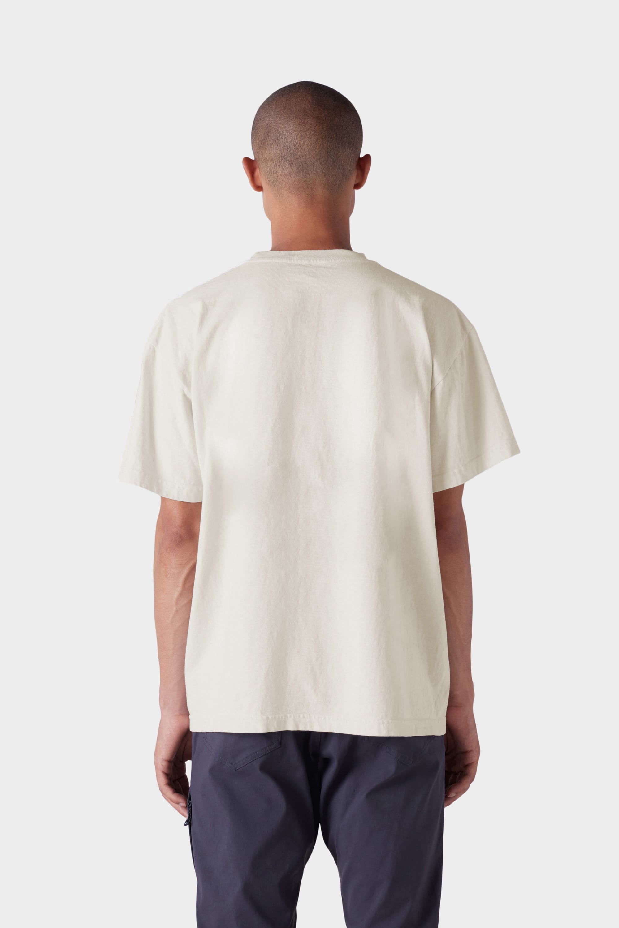 A person with a shaved head stands with their back to the camera, wearing the 686 Mens Bark and Bite Short Sleeve Tee in off-white and dark pants, against a plain light background.