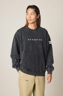 A person with long hair wears a black 686 Men's Mixed Relaxed LS Crewneck and light-colored pants, standing slightly turned against a plain background.