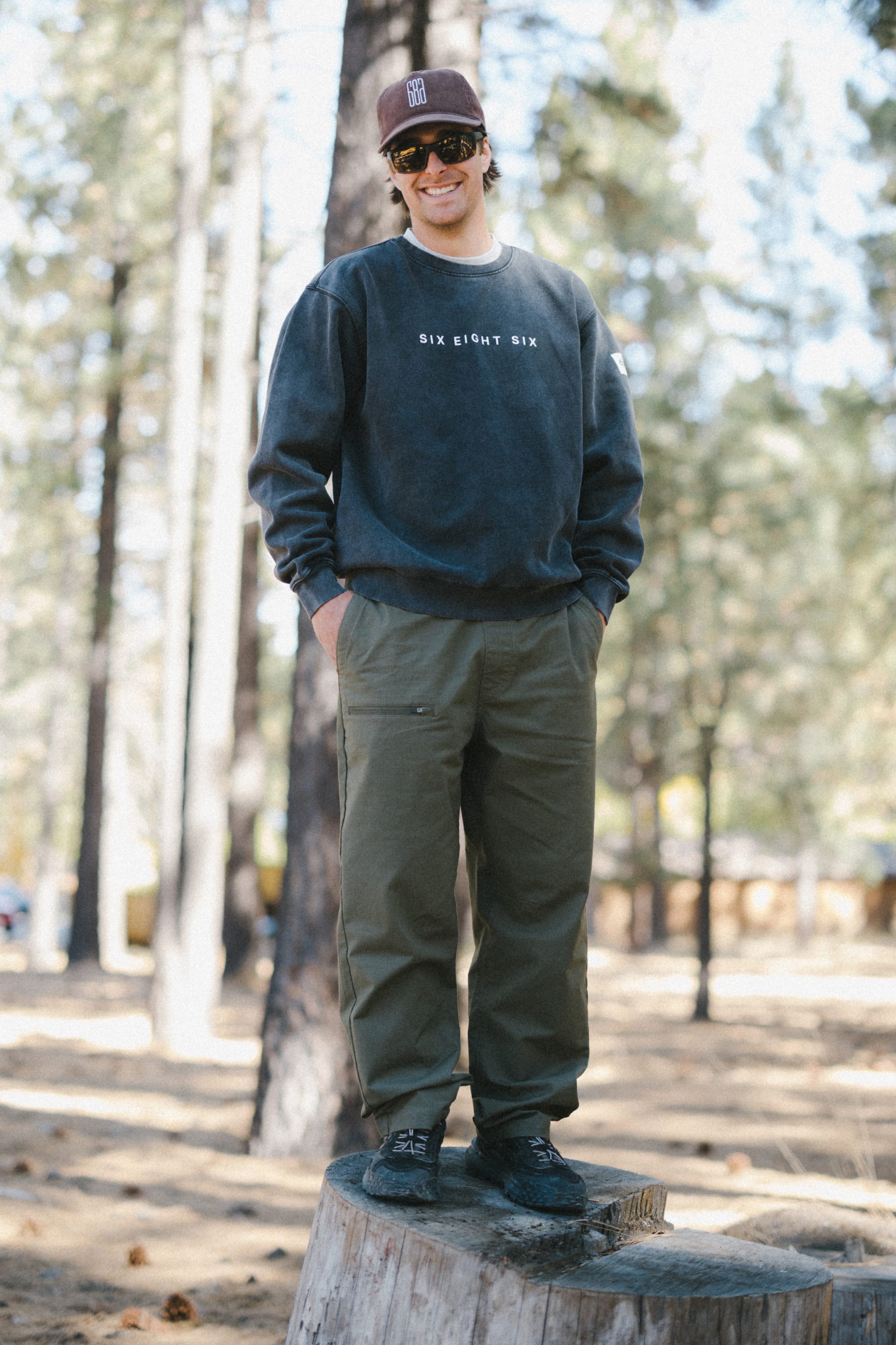 A person wearing sunglasses, a dark 686 Men's Mixed Relaxed LS Crewneck over a classic men's t-shirt, cap, and green pants stands on a tree stump in a forested area, smiling with hands in pockets.
