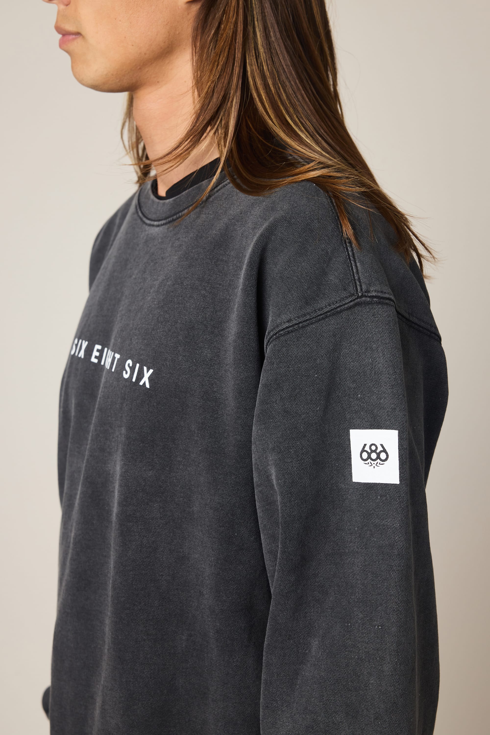 A person with long brown hair is wearing the 686 Men's Mixed Relaxed LS Crewneck, a faded black sweatshirt styled over a classic men's t-shirt. The crewneck features white "SIX EIGHT SIX" text on the chest and a white rectangular "686" patch on the left sleeve. The person is facing left and shown from the chest up.