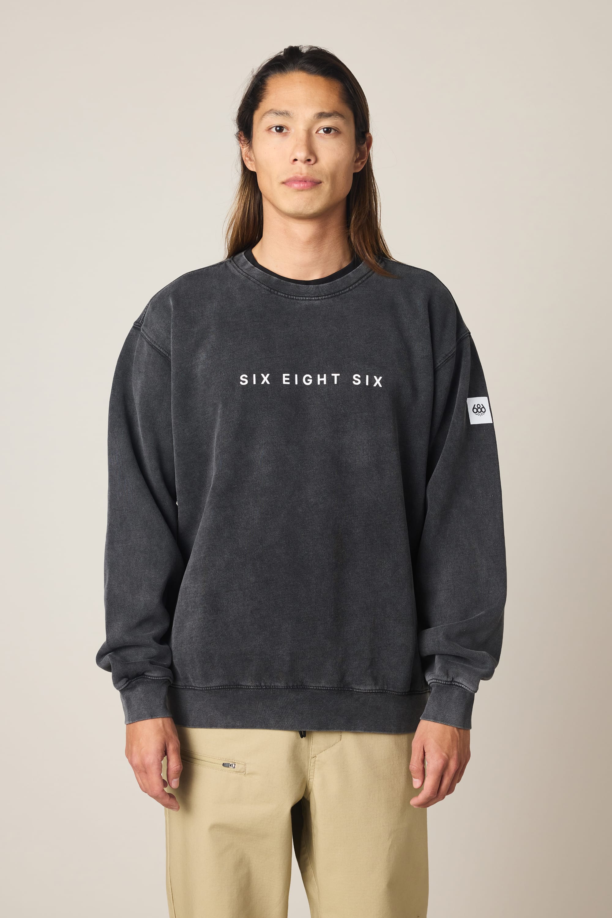 A person with long hair wears the 686 Men's Mixed Relaxed LS Crewneck in black and light khaki pants, standing against a plain, light-colored background.