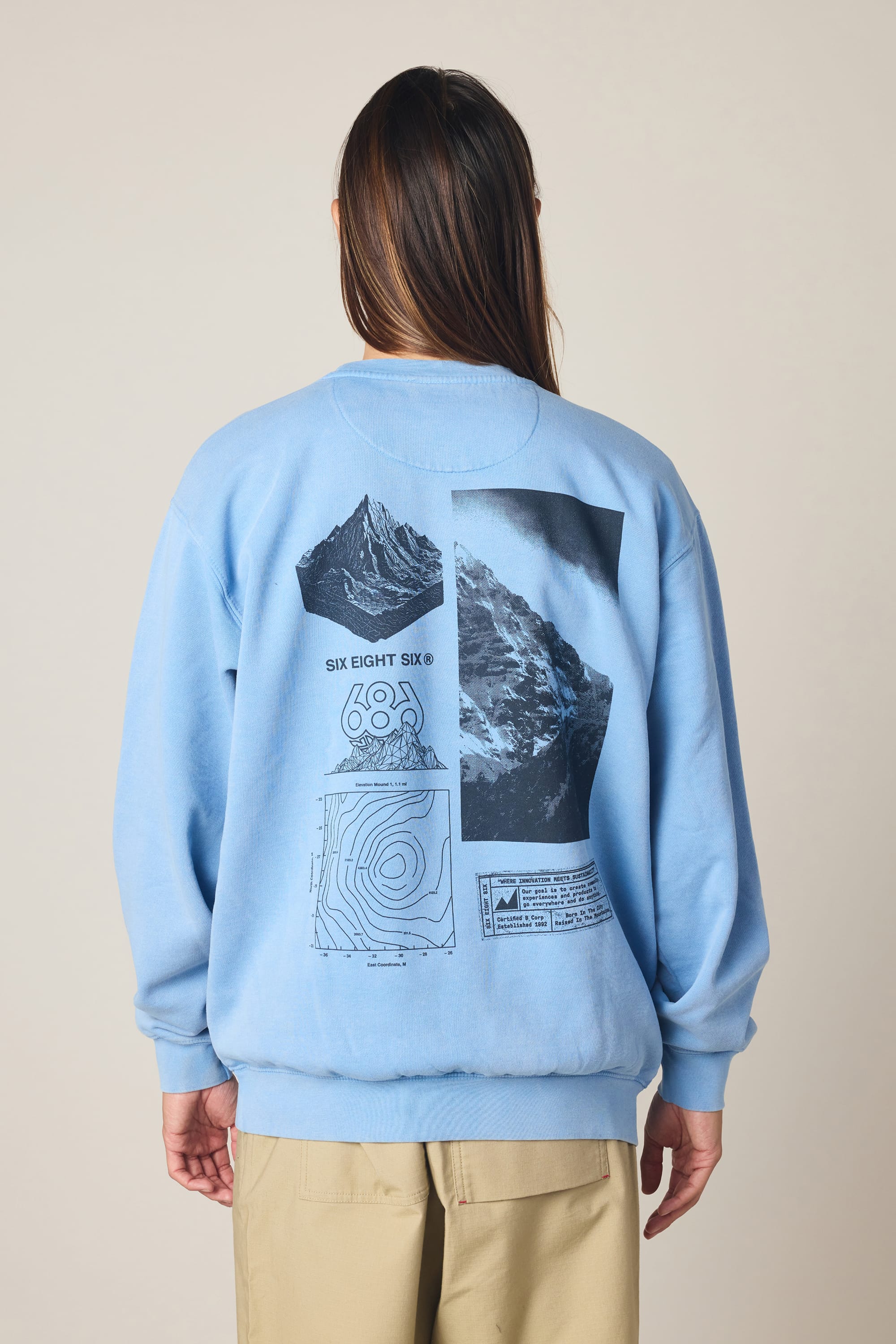 A person stands against a plain background wearing the 686 Men's Mixed Relaxed LS Crewneck by 686—a light blue 100% cotton sweatshirt featuring black mountain graphics, a topographic map, and "SIX EIGHT SIX" printed on the back.