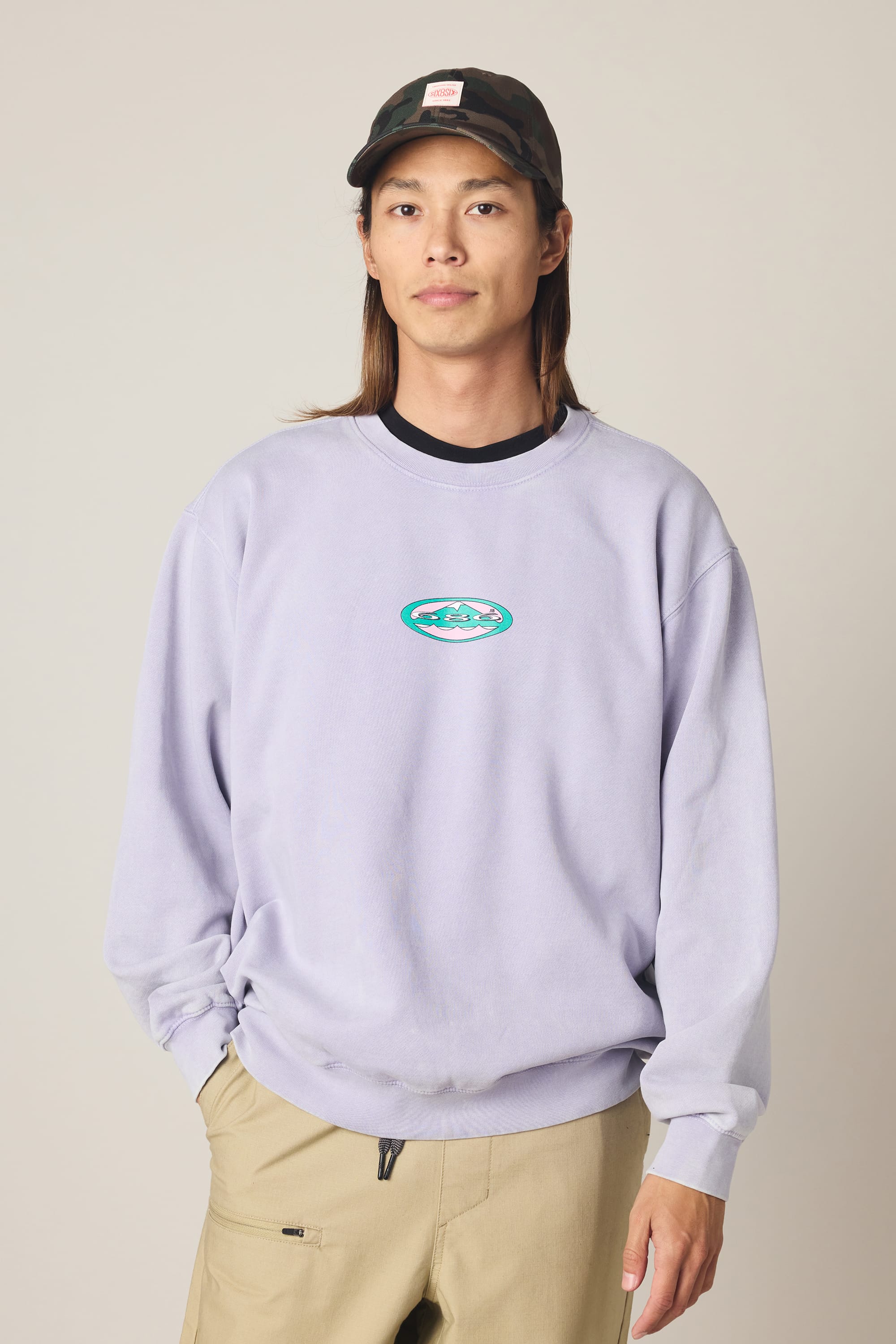 A person with long hair in a black cap, beige pants, and the 686 Men's Mixed Relaxed LS Crewneck by 686—a light purple 100% cotton sweatshirt with a small chest graphic—stands against a neutral background.