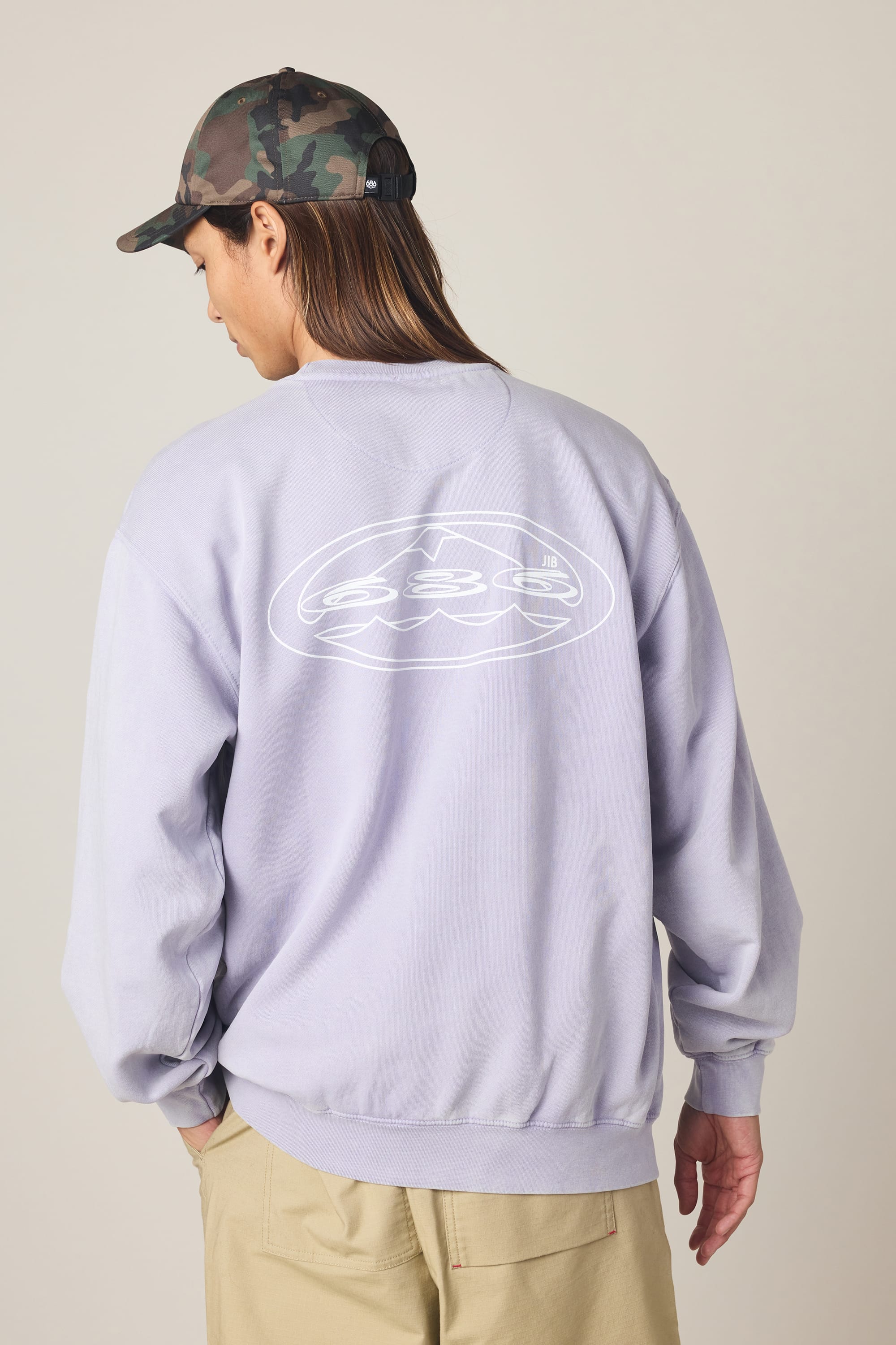 A person wearing the 686 Men's Mixed Relaxed LS Crewneck—a light purple 100% cotton sweatshirt from 686 featuring a white abstract design on the back—paired with tan pants and a camo cap, stands against a plain background.