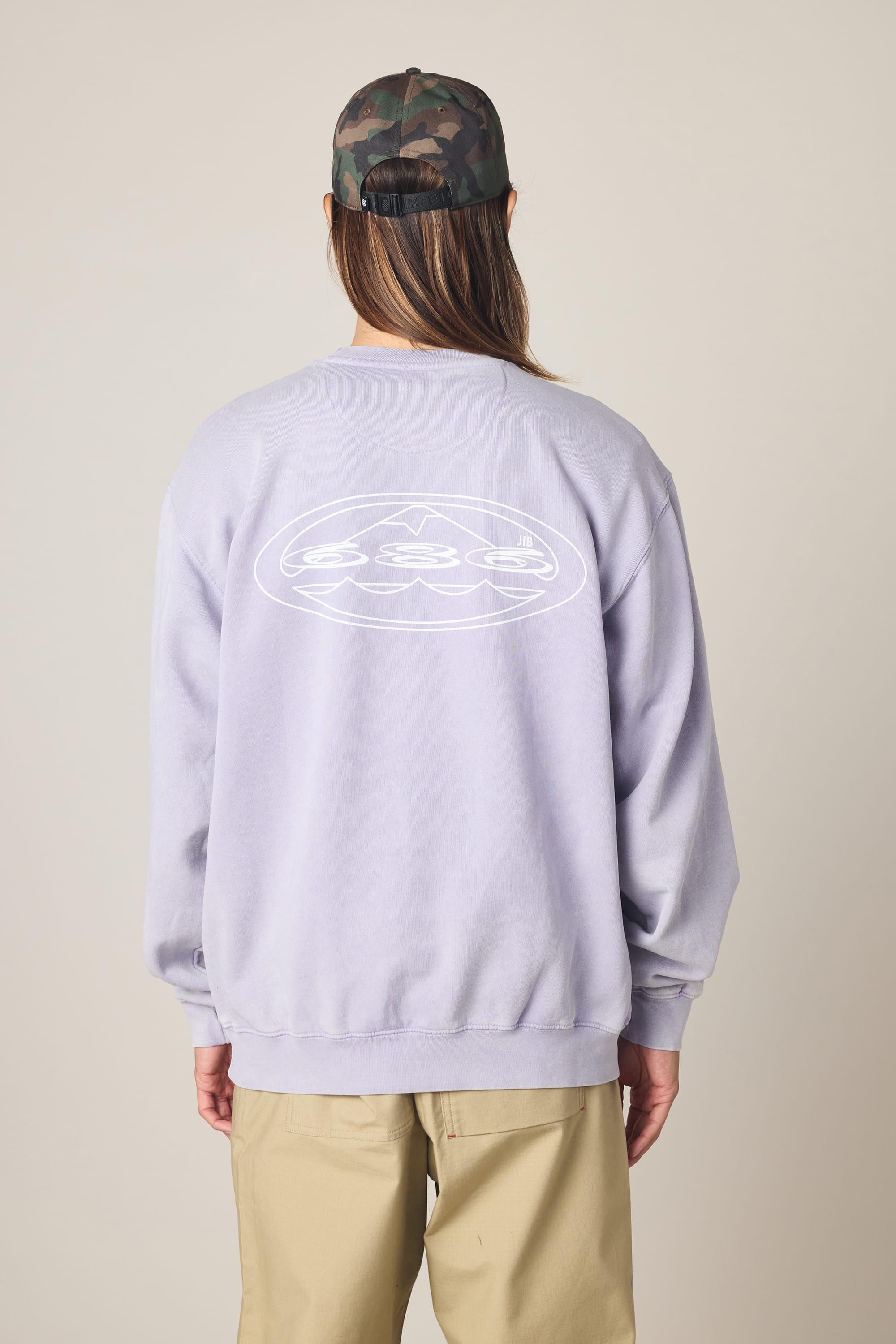 A person faces away from the camera in a 686 Men's Mixed Relaxed LS Crewneck by 686—light purple, 100% cotton, featuring a white oval graphic on the back—paired with beige pants and a green camo cap against a plain background.