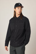 A person with long hair wears a black 686 Men's Whisper Knit Flannel shirt, dark pants, and a black baseball cap with white lettering, standing against a plain light background and looking to the side.
