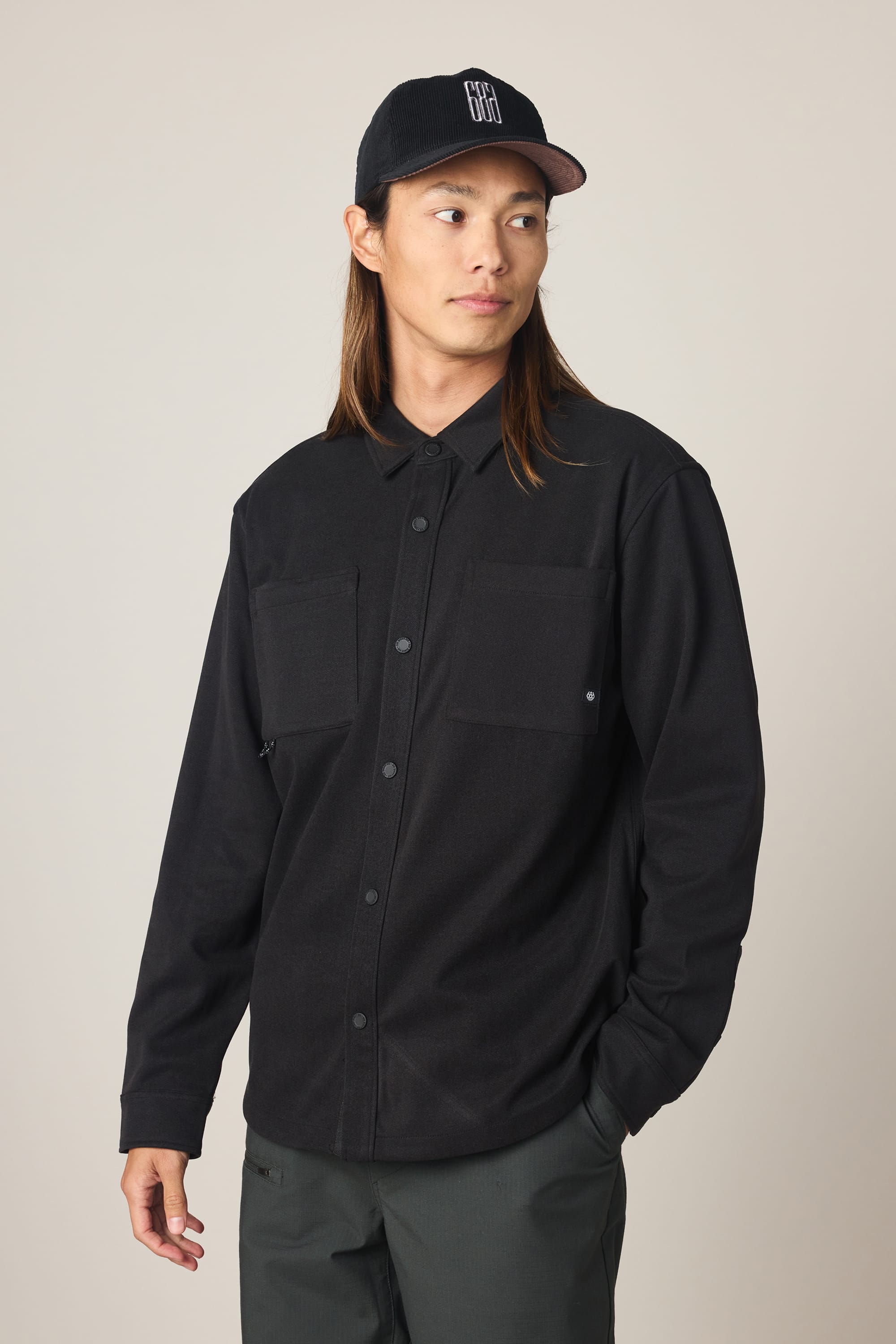 A person with long hair wears a black 686 Men's Whisper Knit Flannel shirt, dark pants, and a black baseball cap with white lettering, standing against a plain light background and looking to the side.