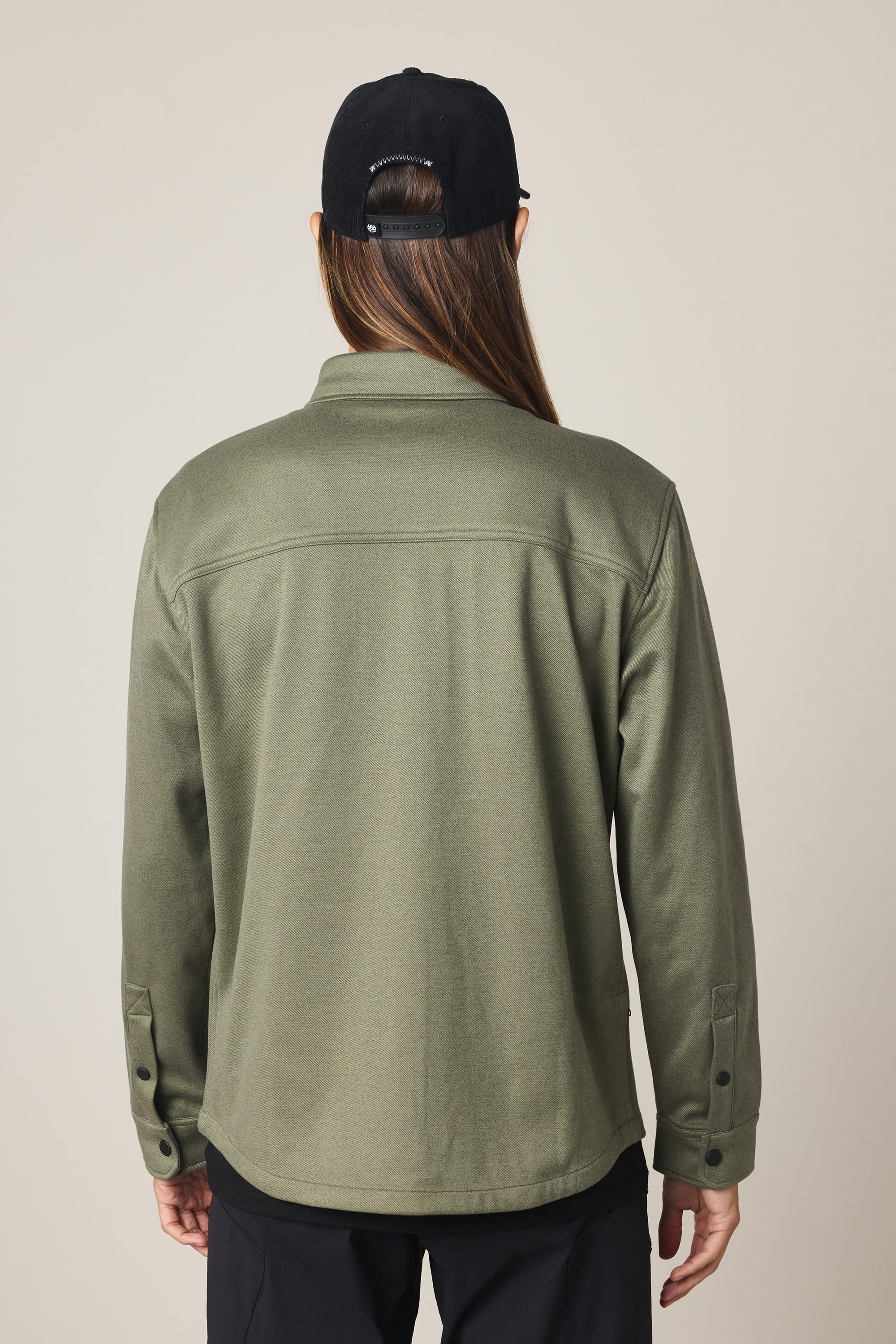 Person with long hair, wearing a black baseball cap and black pants, stands with their back to the camera in an olive green 686 Men's Whisper Knit Flannel shirt by 686 against a plain light background.