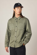 A person with long brown hair, wearing a black baseball cap, black pants, and the 686 Men's Whisper Knit Flannel by 686, featuring two chest pockets in olive green polyester, stands looking to the side with one hand in their pocket against a plain background.