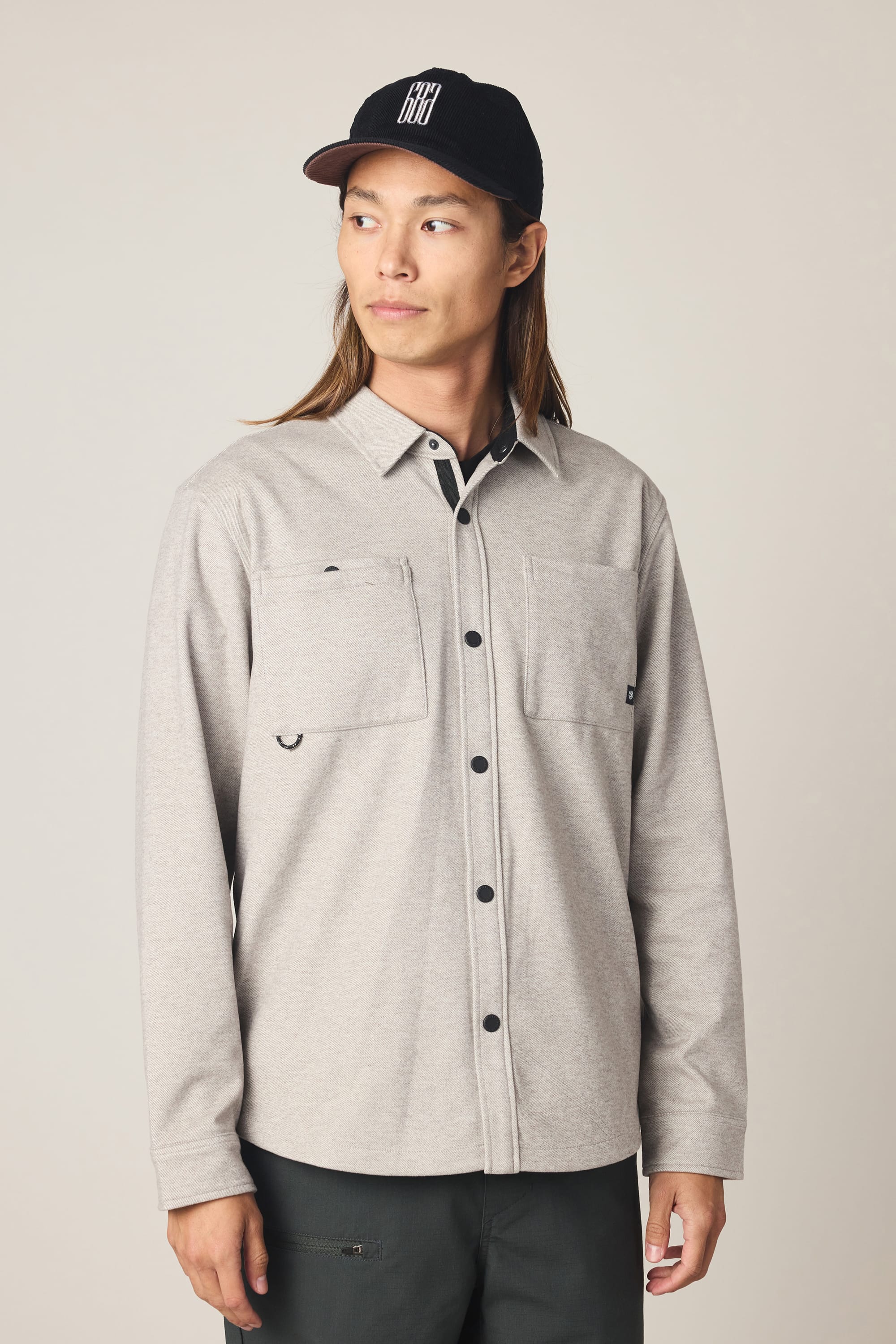 A person with long hair wearing a black cap, dark pants, and the 686 Men's Whisper Knit Flannel by 686 in light gray with front pockets stands facing slightly to the side, looking off-camera.