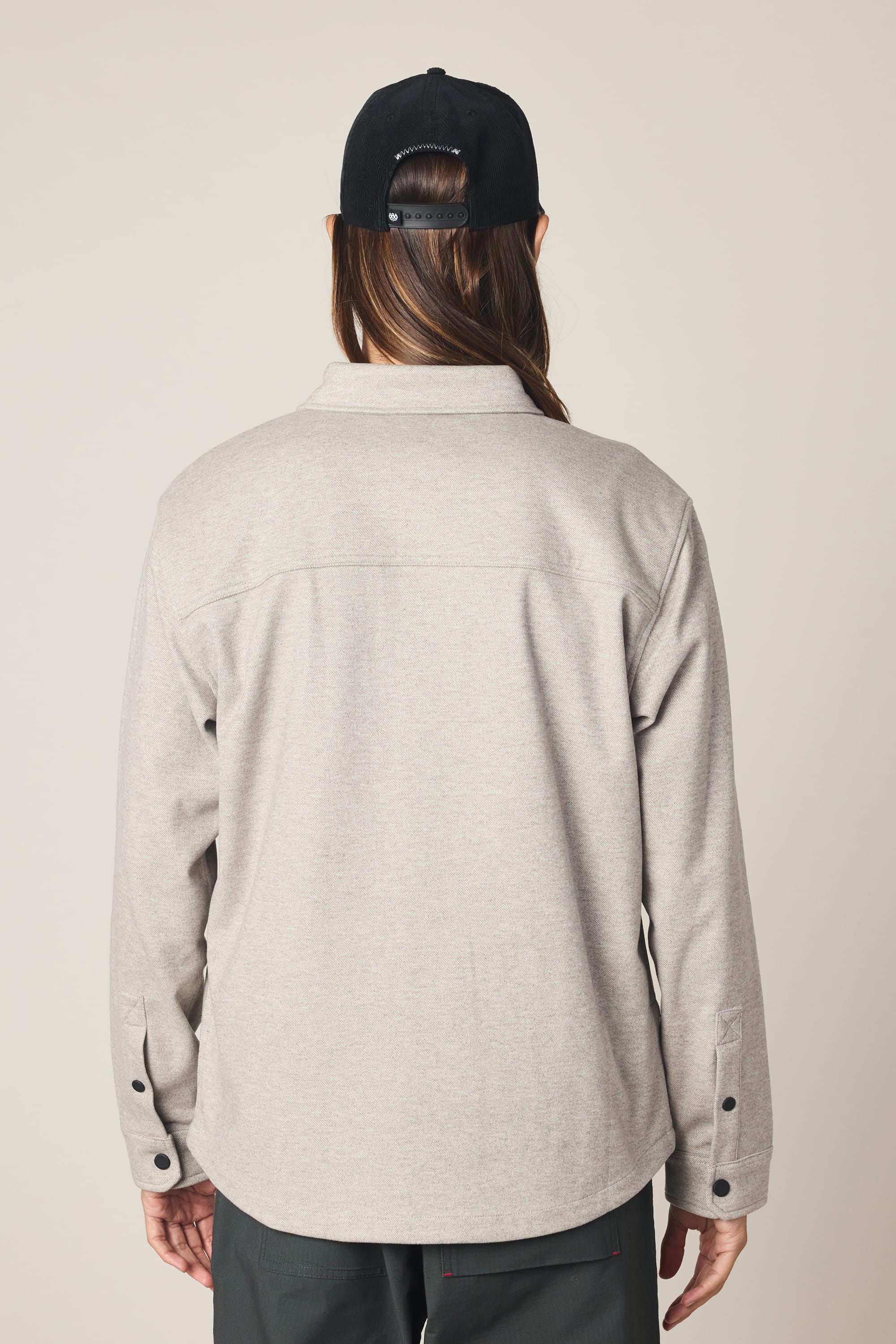 A person with long brown hair, a black backward cap, and dark pants is shown from the back against a plain light background, wearing a light gray 686 Men's Whisper Knit Flannel by 686.