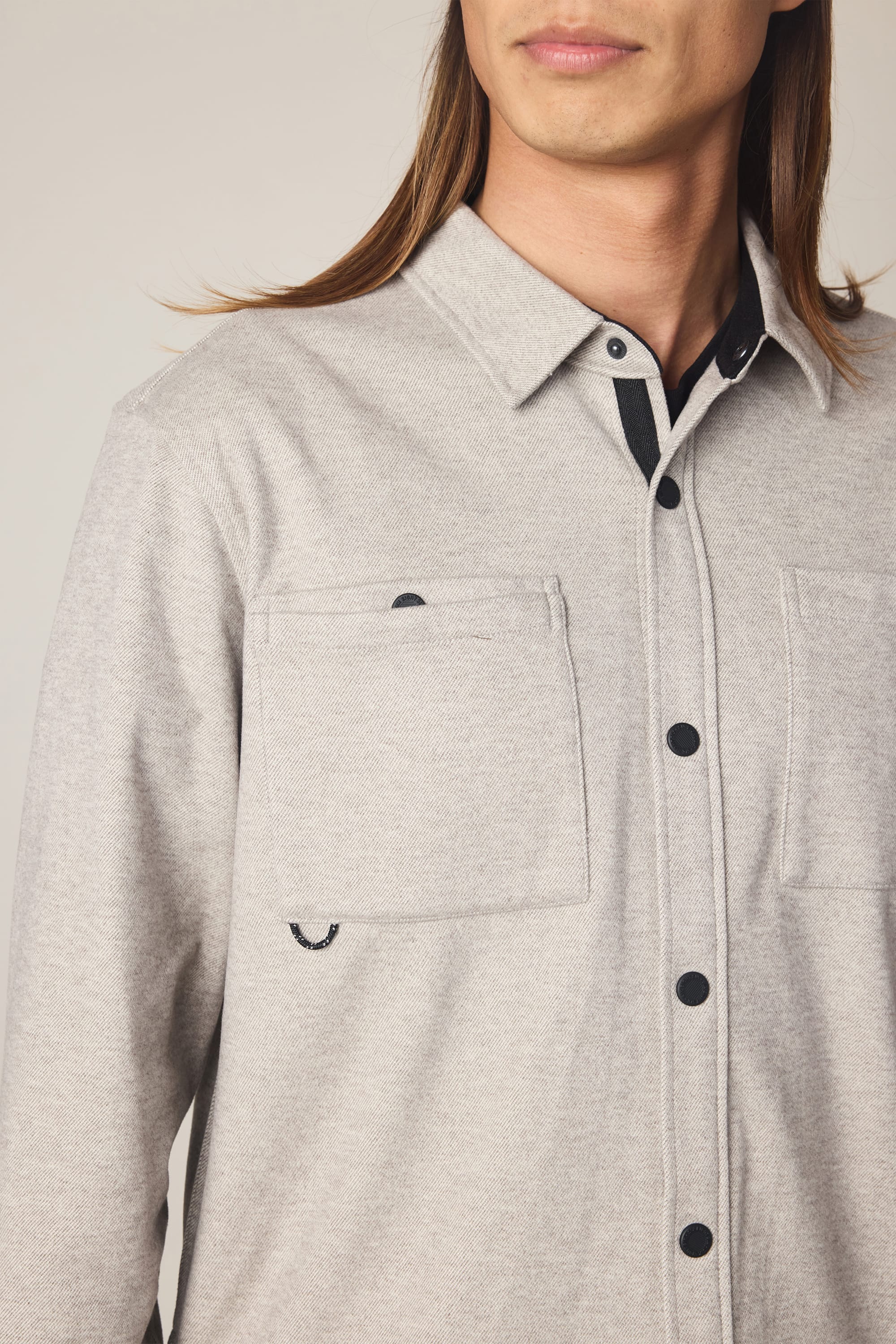 A person with long brown hair is shown from the shoulders to chin, wearing the 686 Men's Whisper Knit Flannel by 686—a light gray polyester shirt with two chest pockets.