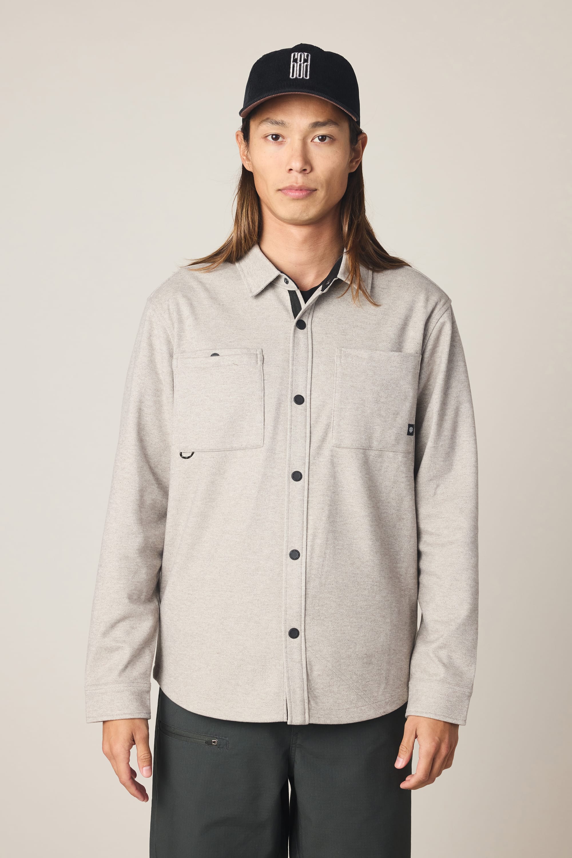 A person with long hair wears a black cap, dark pants, and a light gray 686 Men's Whisper Knit Flannel by 686, featuring two chest pockets, standing against a plain light background.