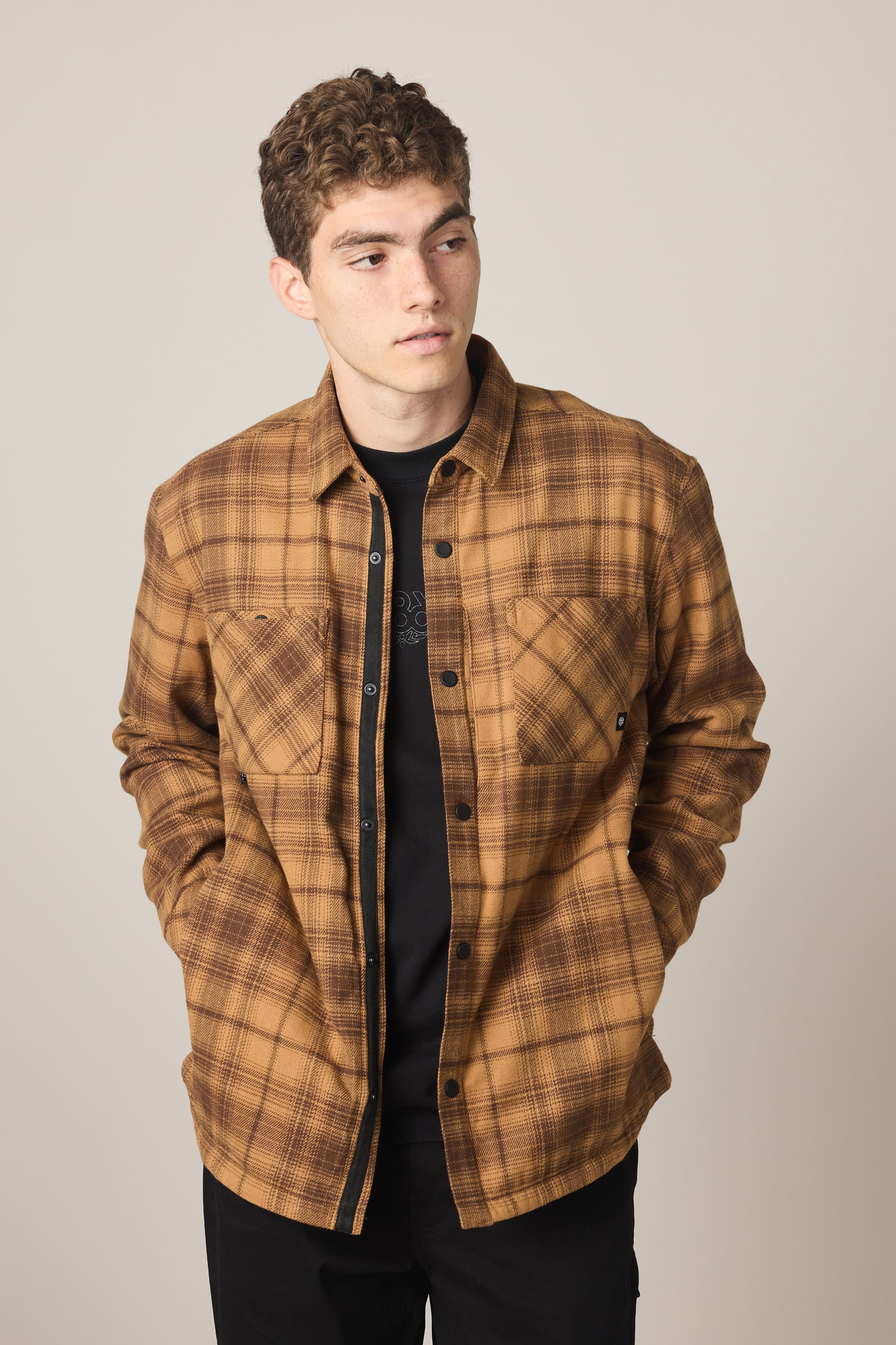 A young man with curly hair wears the 686 Men's Heat Grid Flannel over a black shirt, standing with hands in his pockets and looking to the side against a plain light background.