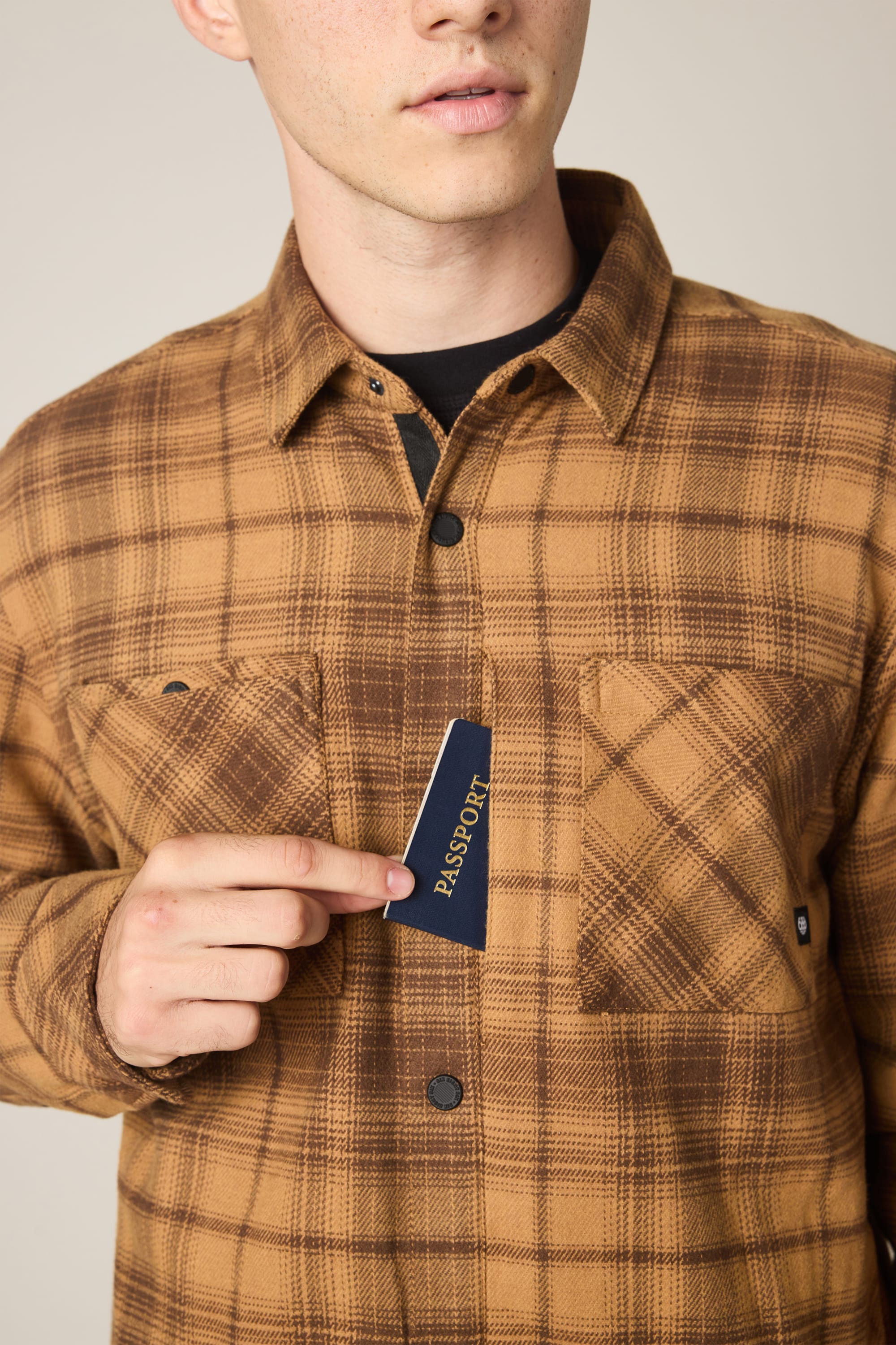 A person wearing the 686 Men's Heat Grid Flannel in brown plaid puts a navy blue passport into the shirt's chest pocket. The person's face is partially visible against a neutral background.