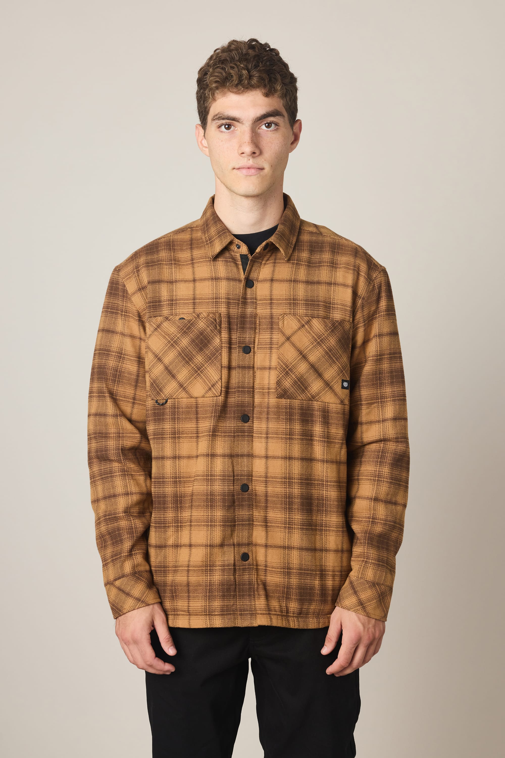 A person wearing the 686 Men's Heat Grid Flannel, a brown plaid polyester shirt with two chest pockets, stands facing forward against a plain light background.