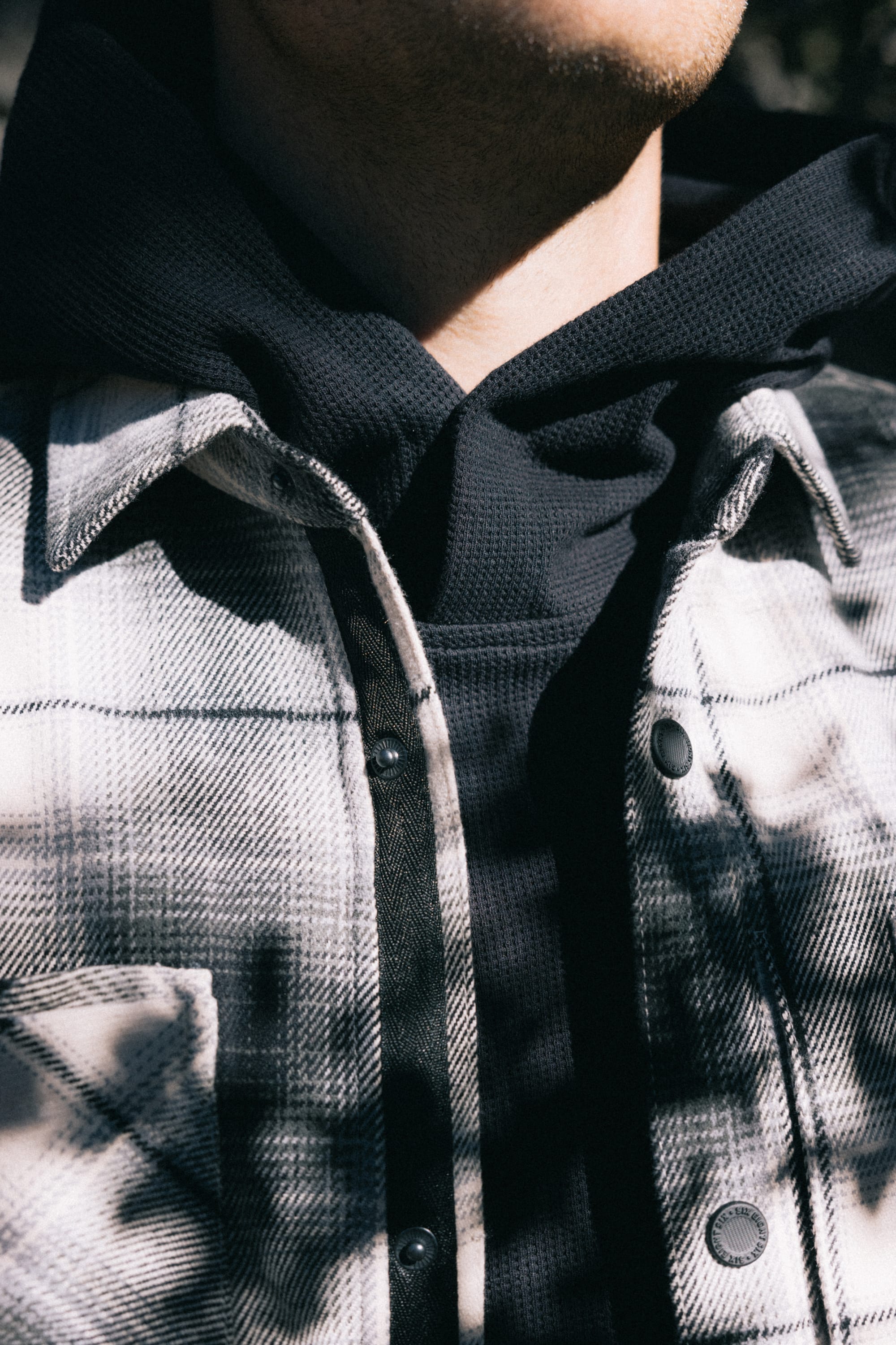A person wearing a black hoodie underneath a white and gray plaid jacket, photographed from the shoulders to the chin in sunlight with shadows across the clothing, showcases a look that's perfectly complemented by the 686 Men's Heat Grid™ Flannel from 686 for added comfort and style.