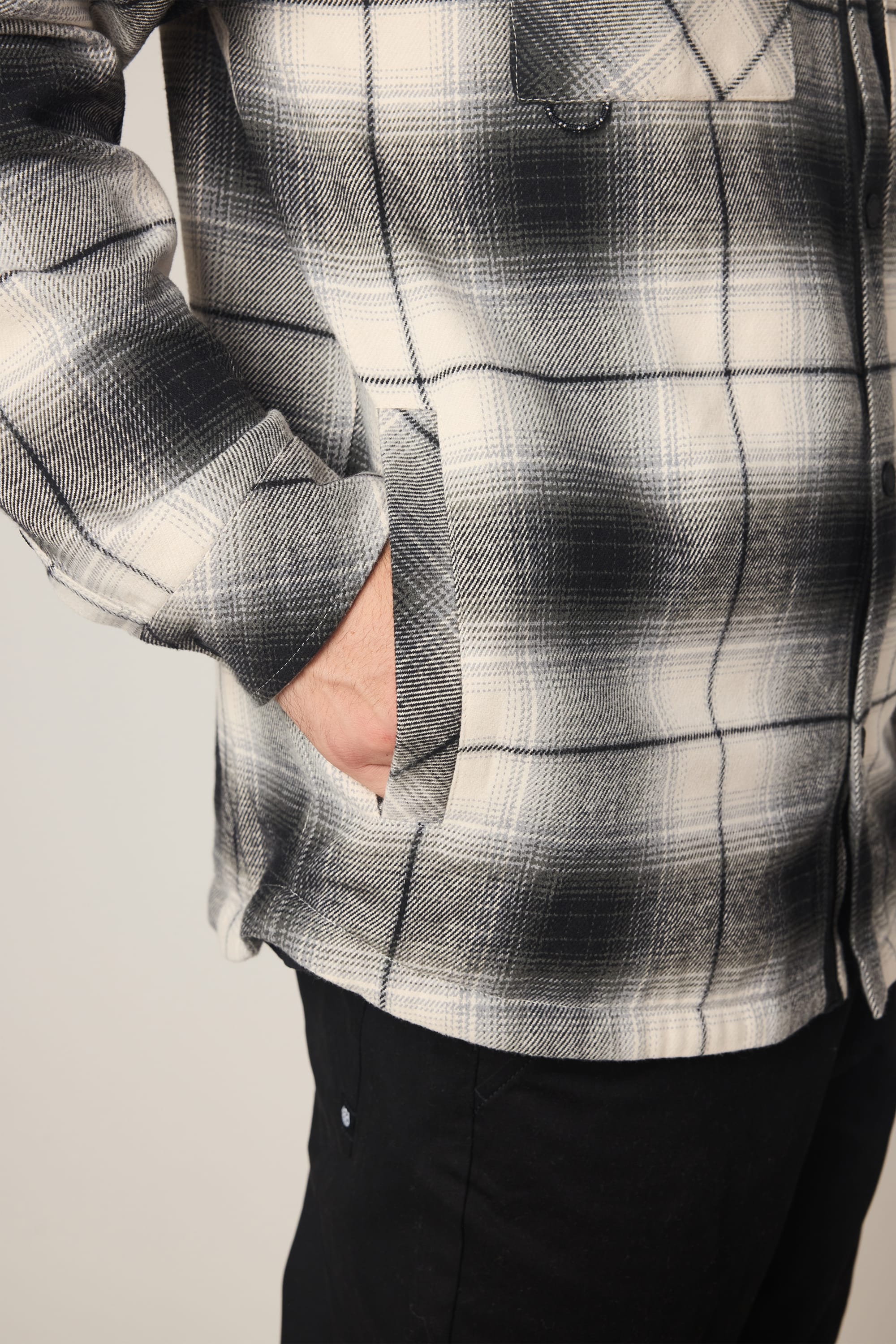 A person wearing the 686 Men's Heat Grid Flannel in black and white plaid with black pants stands with one hand in their pocket. The image highlights the torso and upper legs.