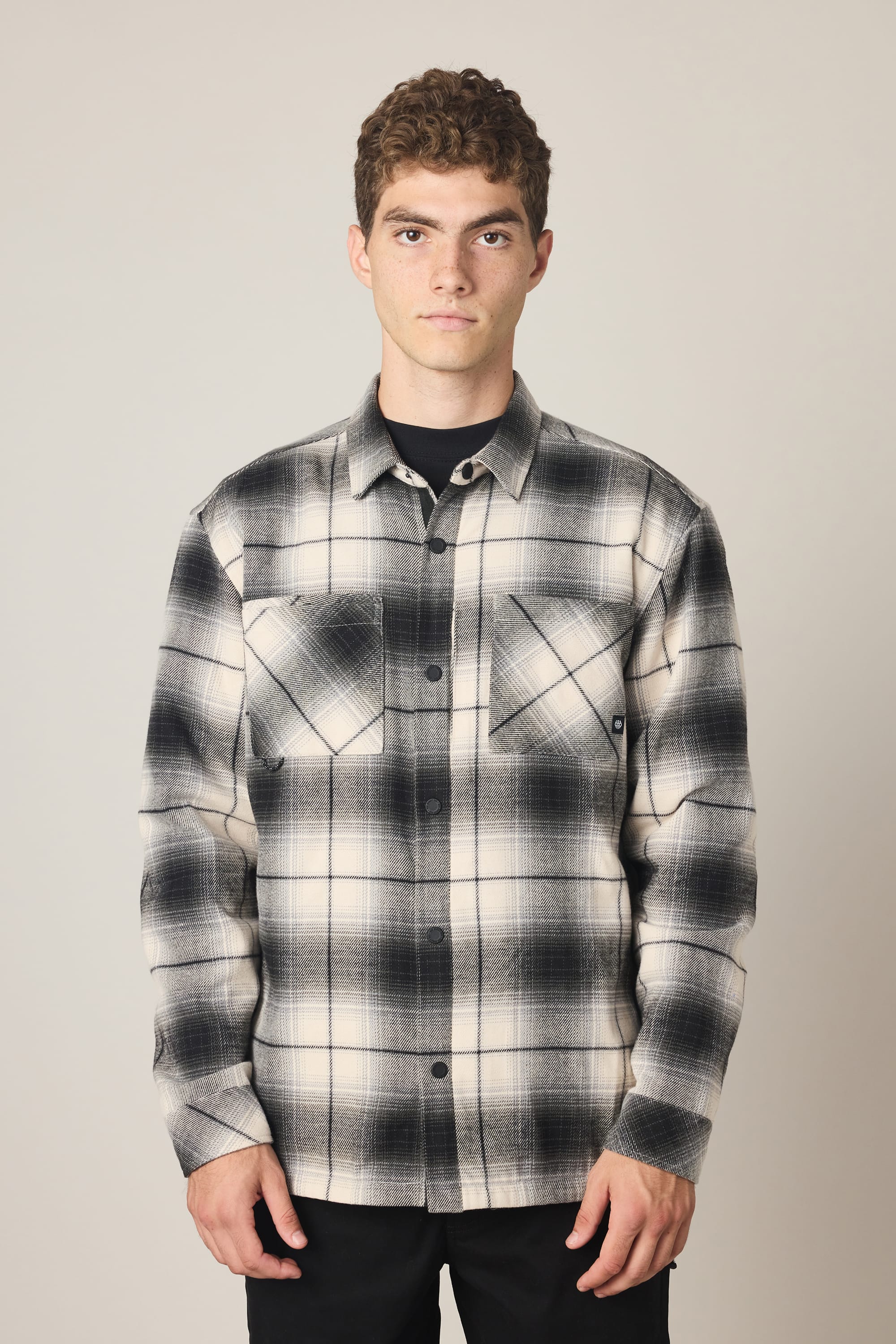 A young man with short curly hair wears the 686 Men's Heat Grid Flannel in black and white plaid over a black t-shirt, standing against a plain light background and looking directly at the camera with a neutral expression.