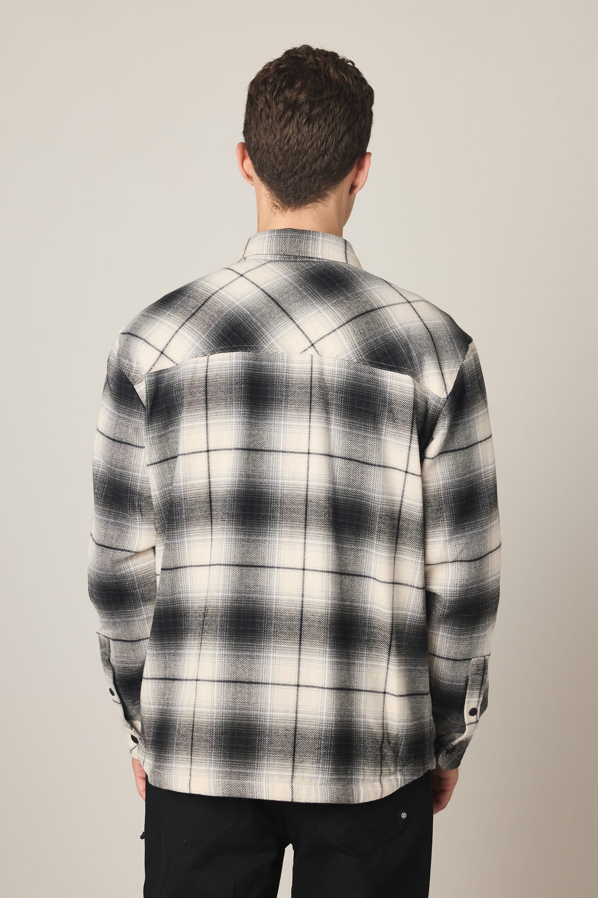 A person with short brown hair, seen from behind, is wearing the 686 Men's Heat Grid™ Flannel by 686—a black and white plaid shirt made from 100% polyester—paired with black pants against a plain light background.