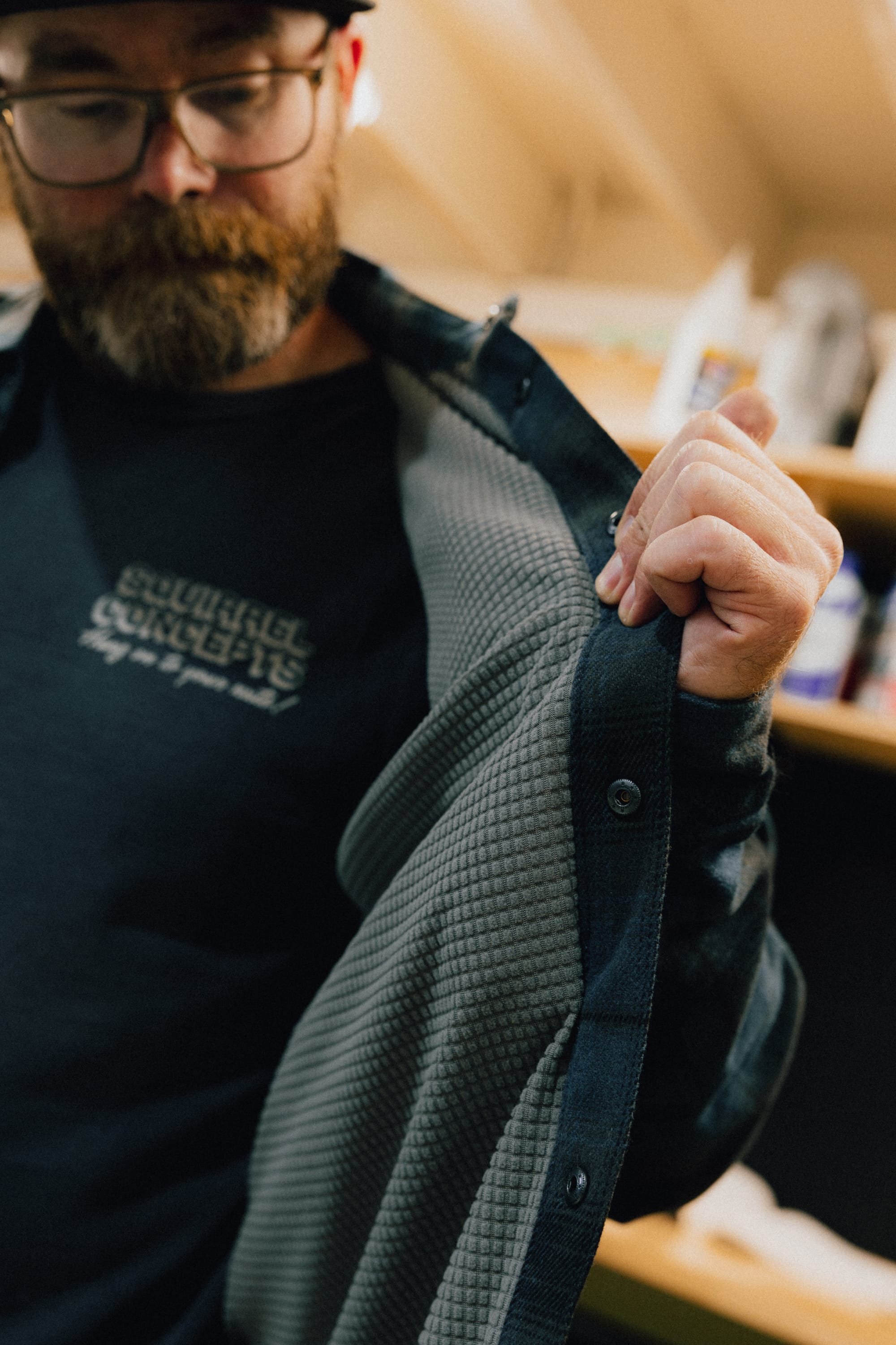 A bearded person wearing glasses opens their 686 Men's Heat Grid™ Flannel shirt, revealing its textured gray inner lining. Shelves with various items can be seen in the blurred background.