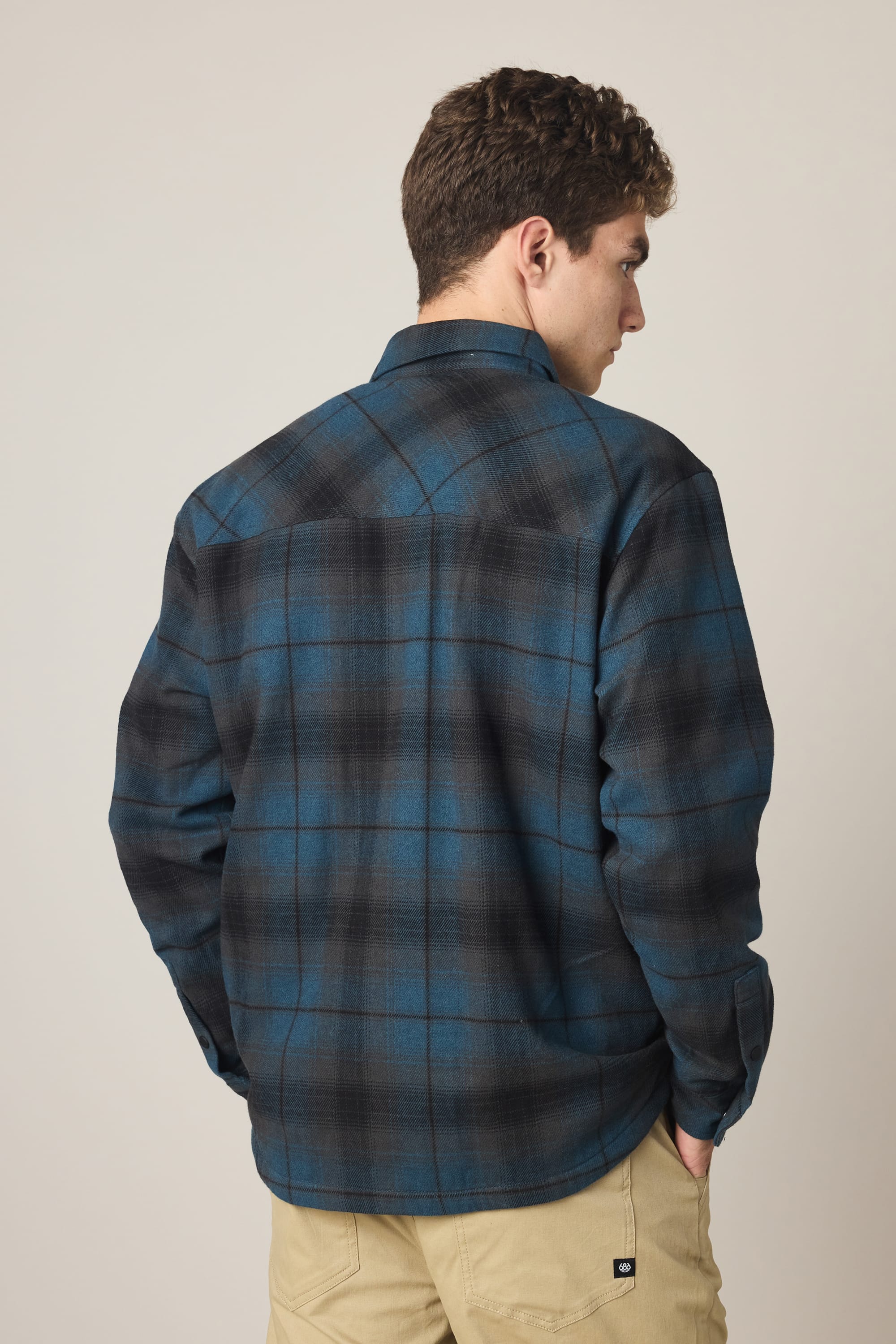 A person with short curly hair, seen from the back, wears a blue and black 686 Men's Heat Grid Flannel shirt and beige pants against a plain light background.