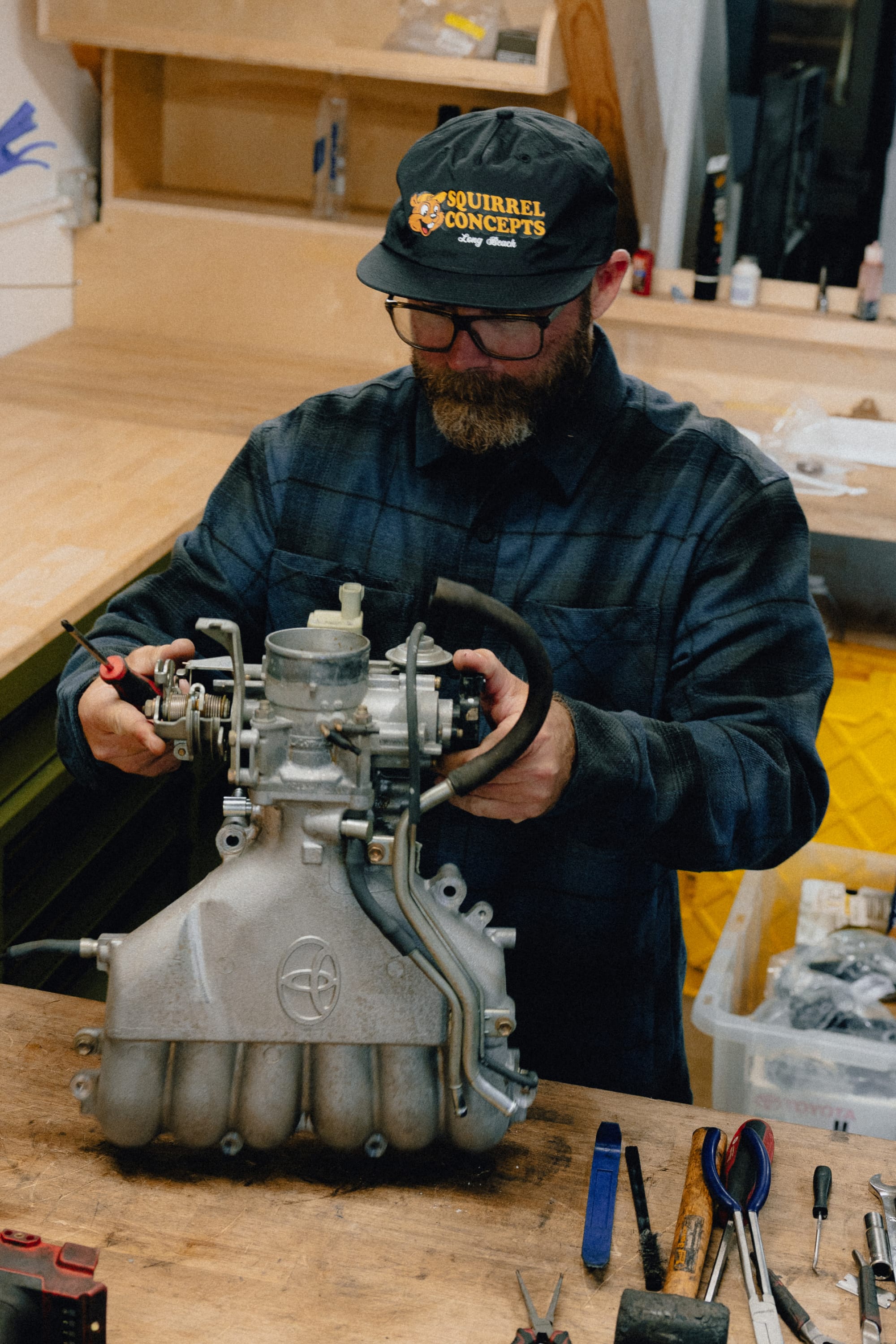A bearded person wearing glasses and a cap, dressed in a comfortable 686 Men's Heat Grid™ Flannel by 686, works on a metal engine part at a wooden workbench, surrounded by tools in a workshop.