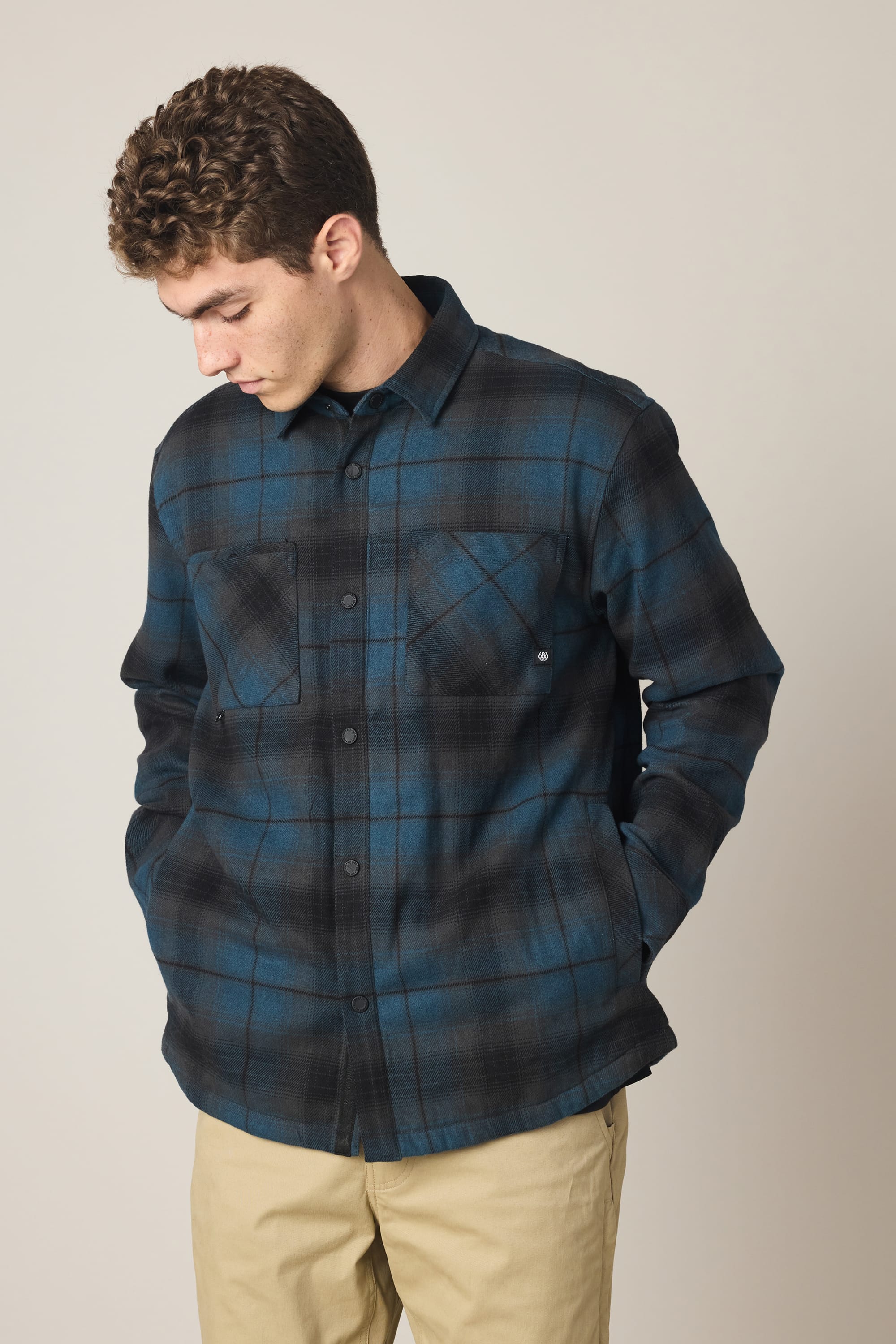 A man with short curly hair looks down while wearing the 686 Men's Heat Grid Flannel in blue and black plaid with front pockets, paired with tan pants, against a plain light background.
