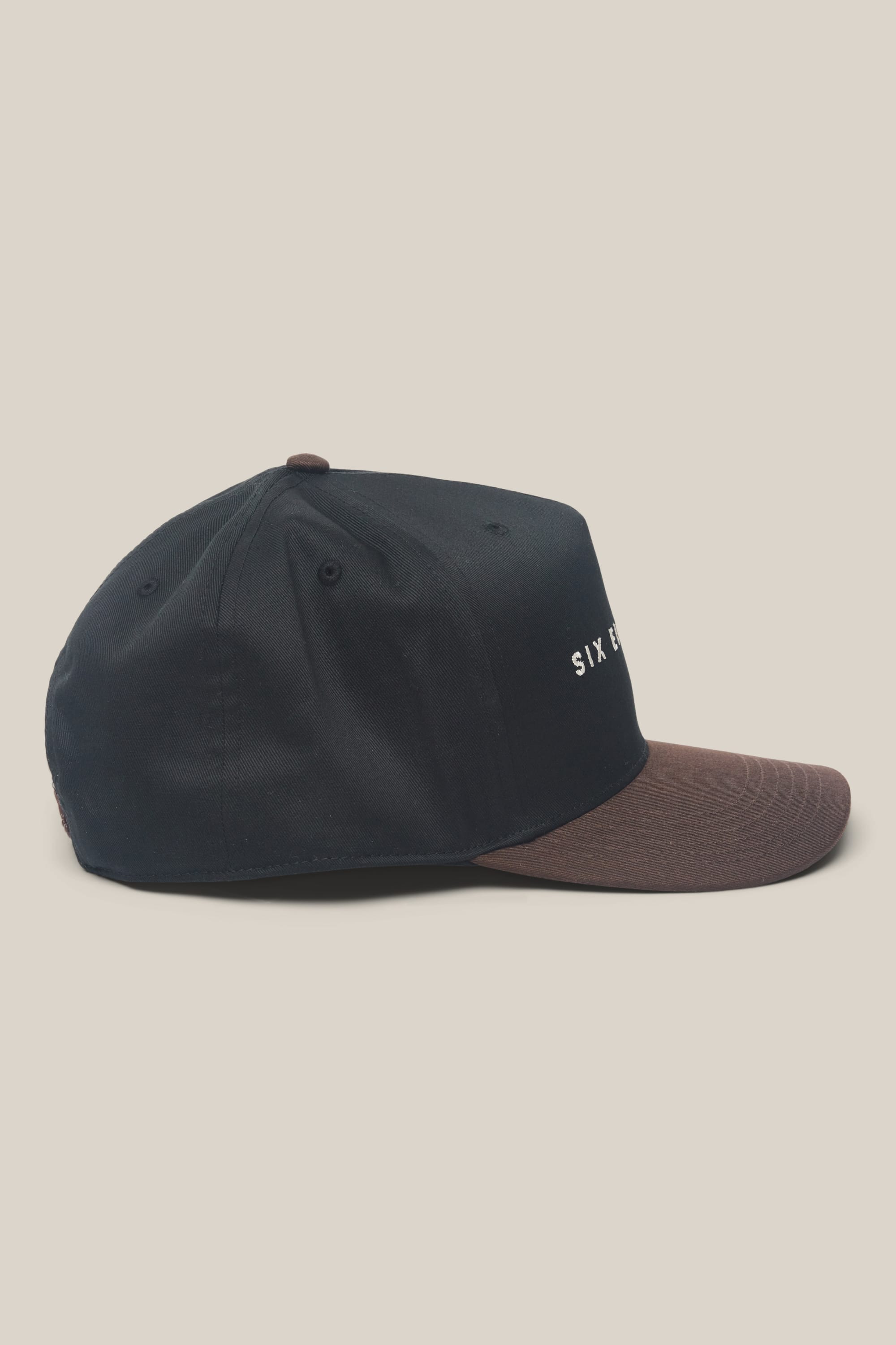 The 686 High-Pro Ses Two Tone Structured 5 Panel Hat by 686 is a black men's hat with a brown brim, shown from the side. Made from 100% cotton, it displays partially visible white text on the front panel against a plain cream background.
