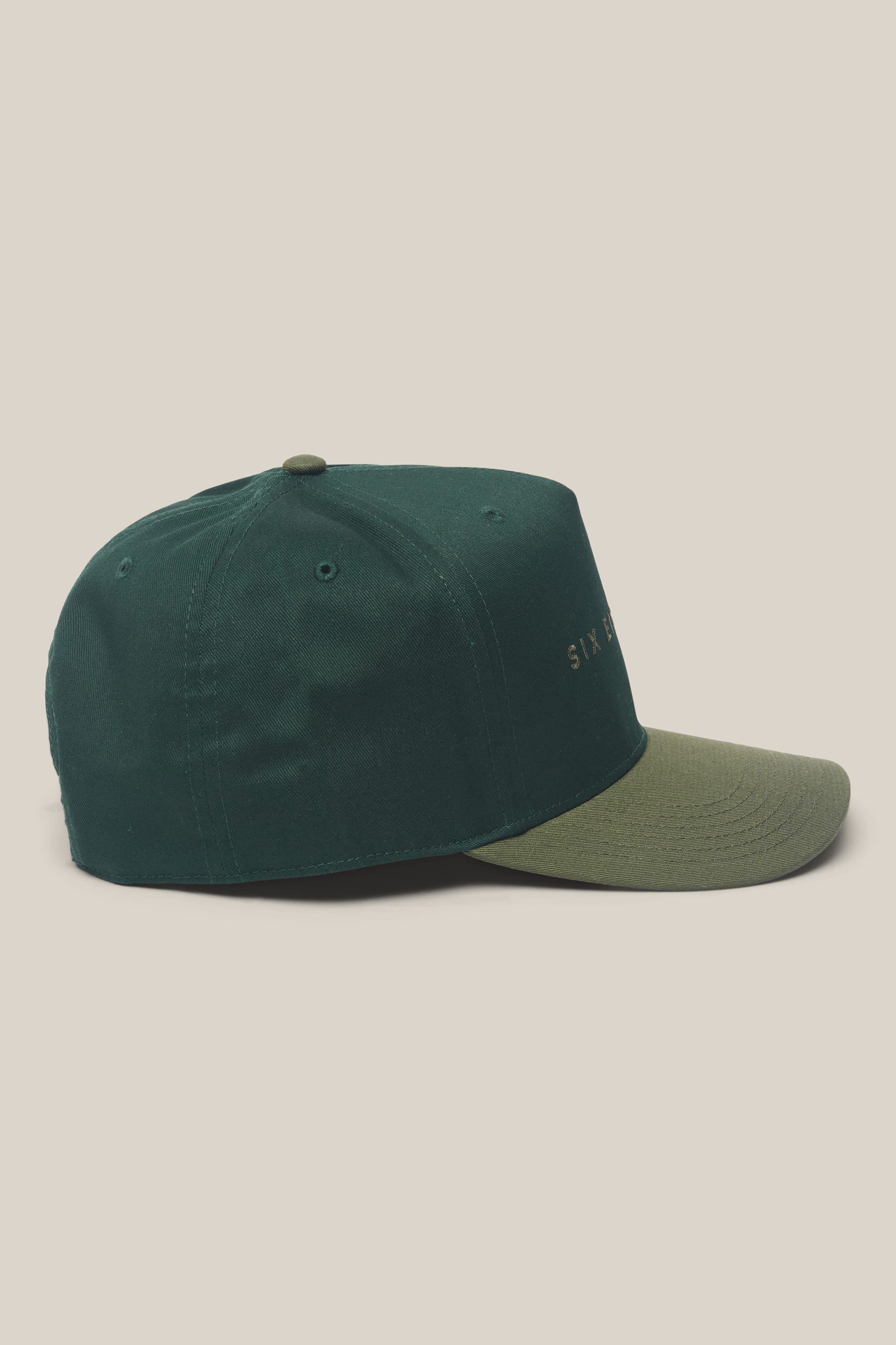 Dark green 686 High-Pro Ses Two Tone Structured 5 Panel Hat with an olive green brim, displayed in side view on a light beige background. This men's hat by 686 features small gold text on the front, partially visible, reading SIX B.