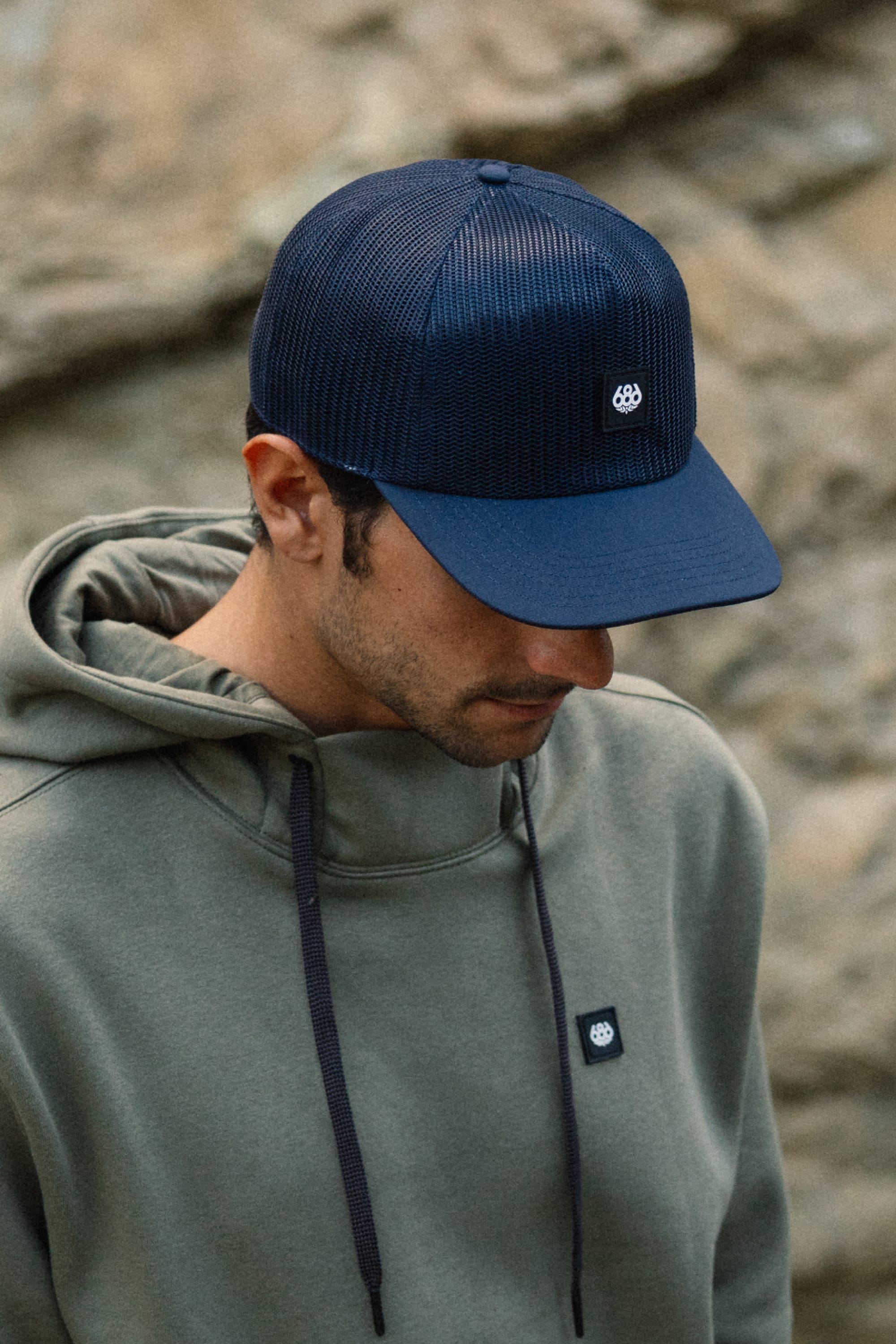 Wearing the 686 High-Pro Knockout Structured 5 Panel Hat by 686 in dark blue cotton, along with an olive green hoodie featuring matching black-and-white logo patches, a person looks down outdoors against a rocky backdrop.
