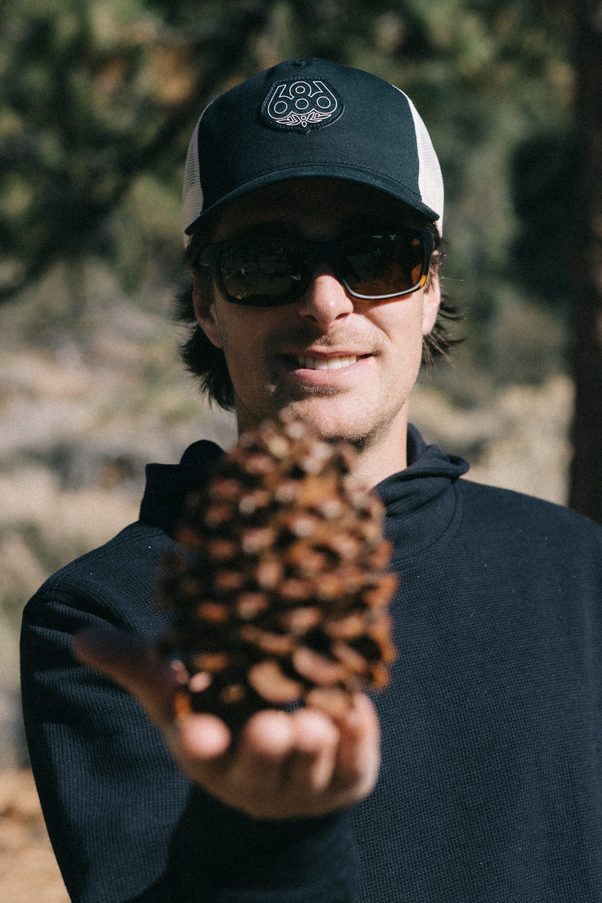 A person wearing sunglasses and a 686 Mid-Pro Wreath Unstructured 5 Panel Mesh Hat by 686 holds a large pine cone toward the camera, with trees and sunlight blurred in the background.