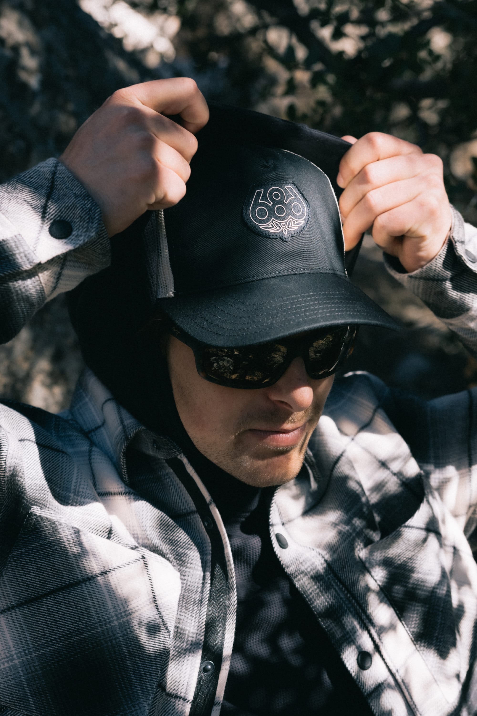 A person wearing sunglasses and a plaid shirt adjusts a 686 Mid-Pro Wreath Unstructured 5 Panel Mesh Hat by 686 outdoors in dappled sunlight.