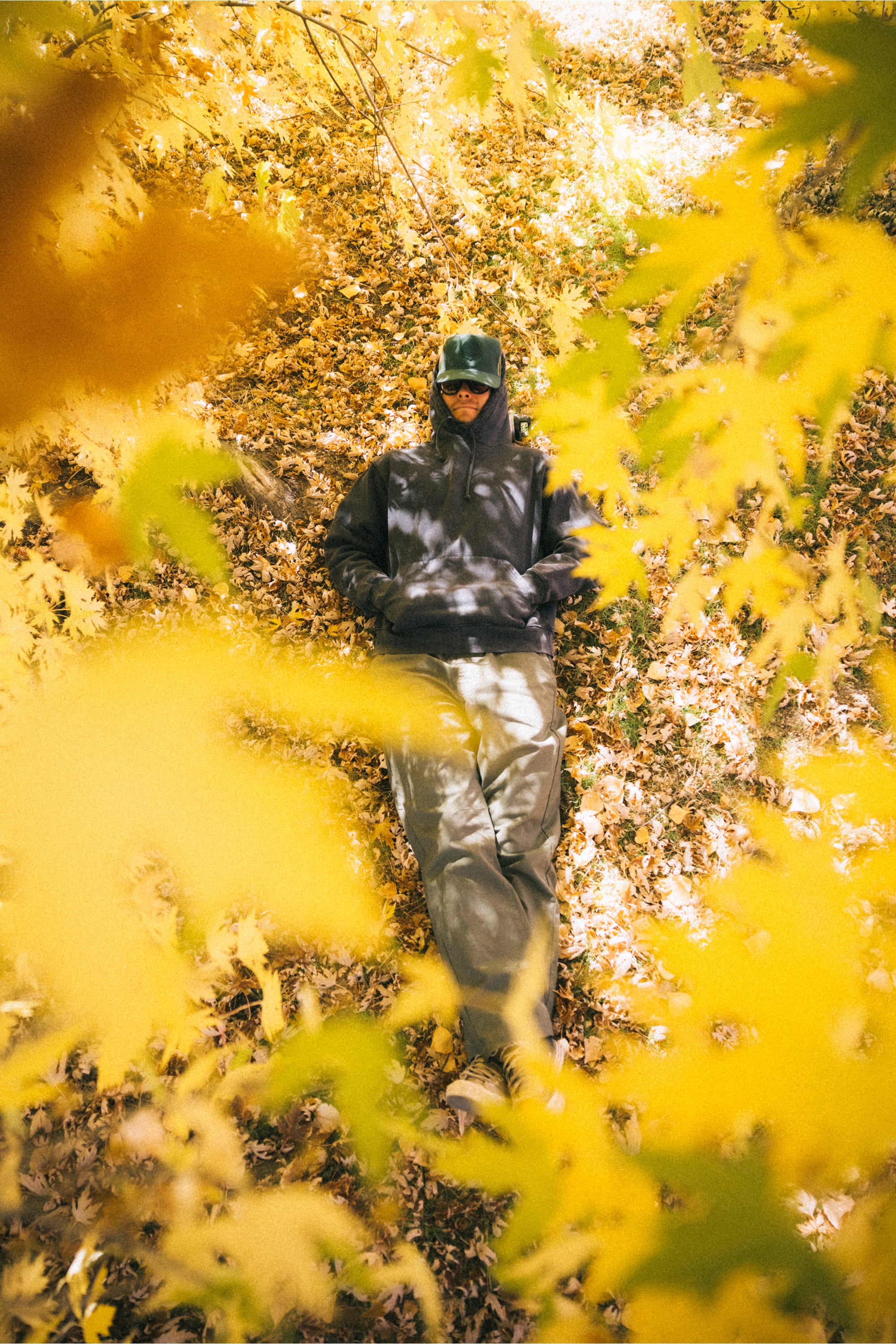 A person wearing sunglasses, a hoodie, and the 686 Mid-Pro Wreath Unstructured 5 Panel Mesh Hat by 686 lies on fallen leaves, surrounded by vibrant yellow autumn foliage as sunlight filters through the branches above.