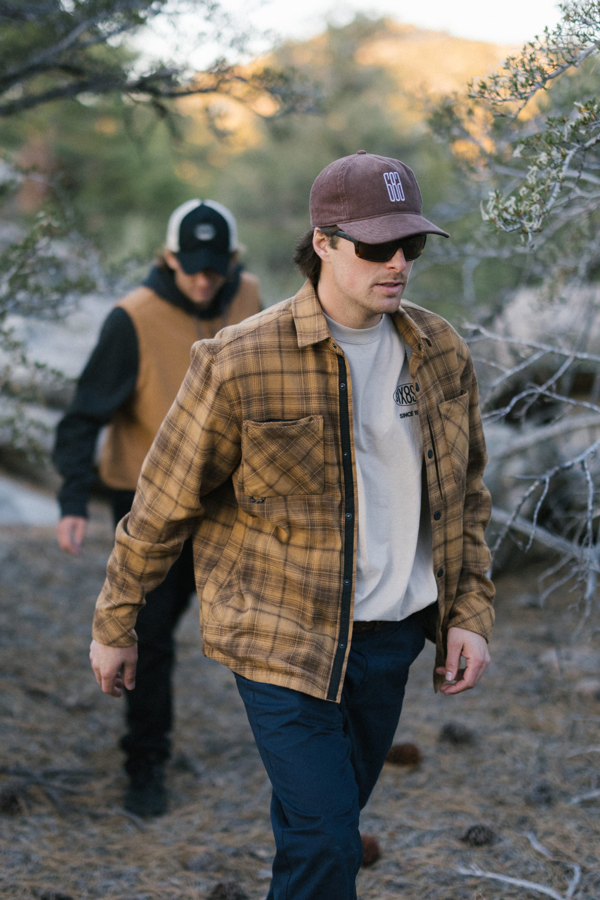 Two men walk outdoors through a wooded area. The man in front wears a brown plaid jacket, sunglasses, and a men's hat. The man behind sports the 686 Mid-Pro Ghost Cord Unstructured 5 Panel Hat by 686, along with a tan vest and black sleeves. Both appear to be hiking.