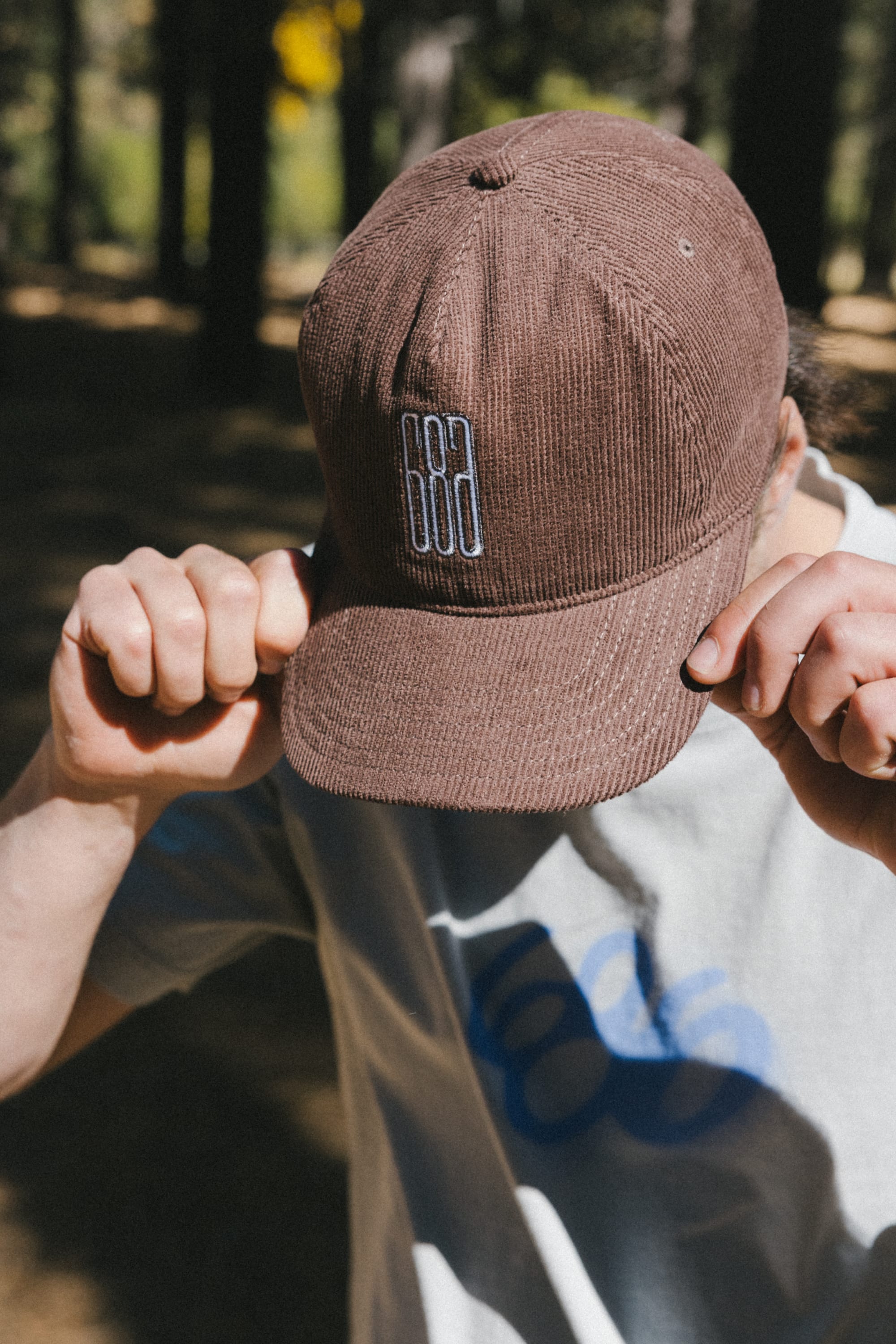 A person outdoors tilts their 686 Mid-Pro Ghost Cord Unstructured 5 Panel Hat down, obscuring their face. They wear a light-colored shirt with a blue design, while blurred trees and sunlight fill the background.