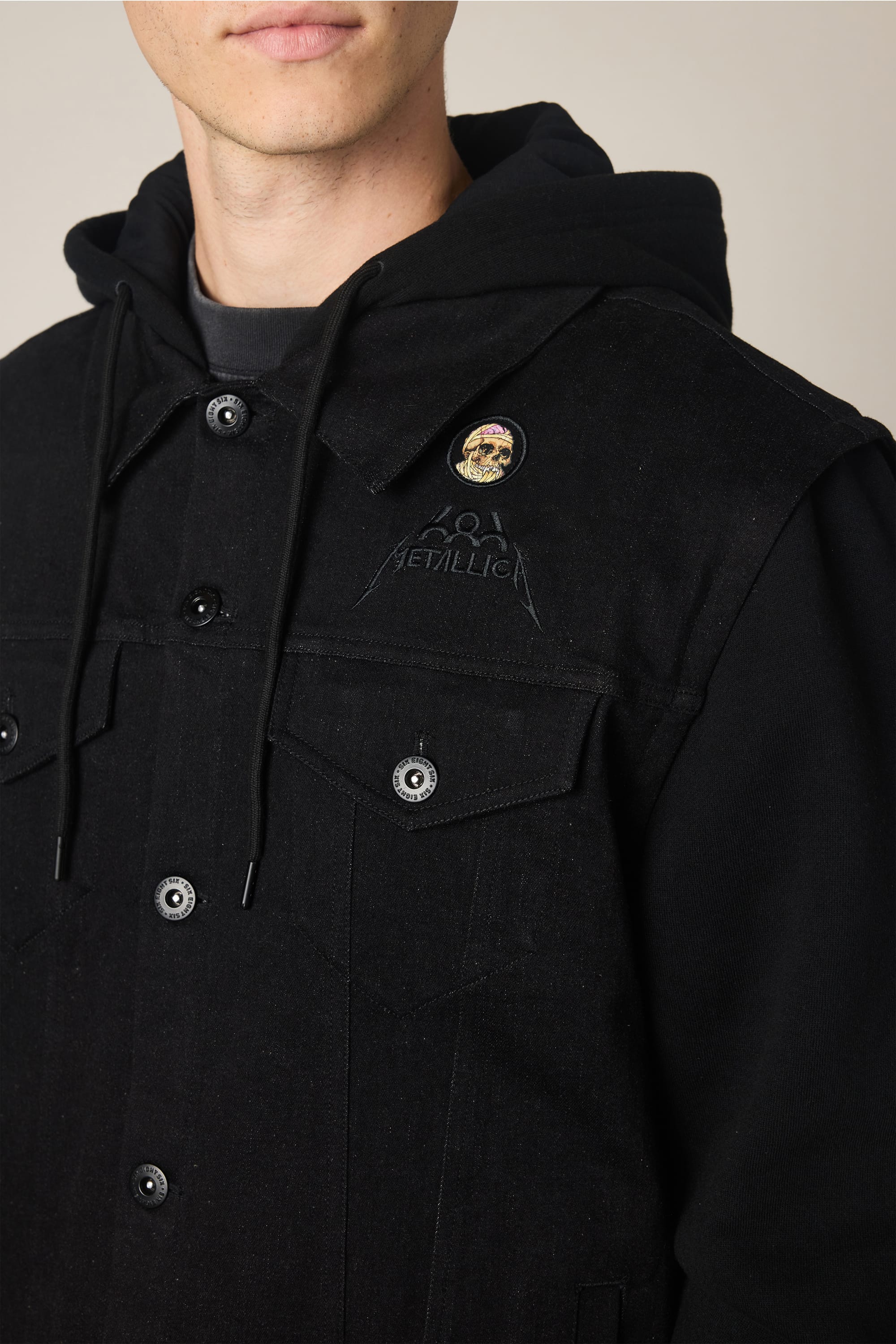 A person wears the 686 Men's Metalhead Trucker Jacket, a black waterproof piece with button details and a Metallica logo on the chest. A colorful round pin is attached to the left side. The person's face is partially visible.
