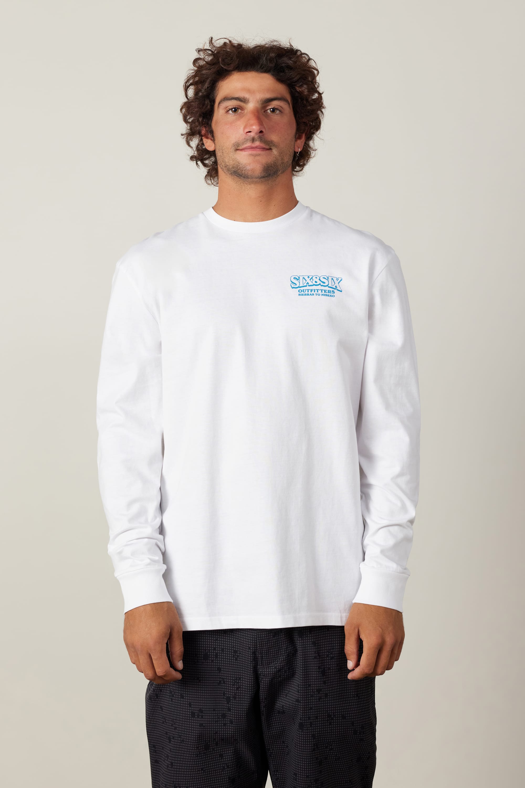 A man with curly hair wears a white 686 Fitted Authentic L/S Tee featuring a small blue and aqua STUSSY DESIGNS logo on the left chest, paired with black shorts, standing against a plain light background.