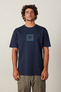 A man with curly hair faces forward wearing the 686 Knockout Authentic S/S Tee in navy blue, featuring a small 686 logo on the chest, paired with olive green patterned shorts. The background is plain and light-colored.