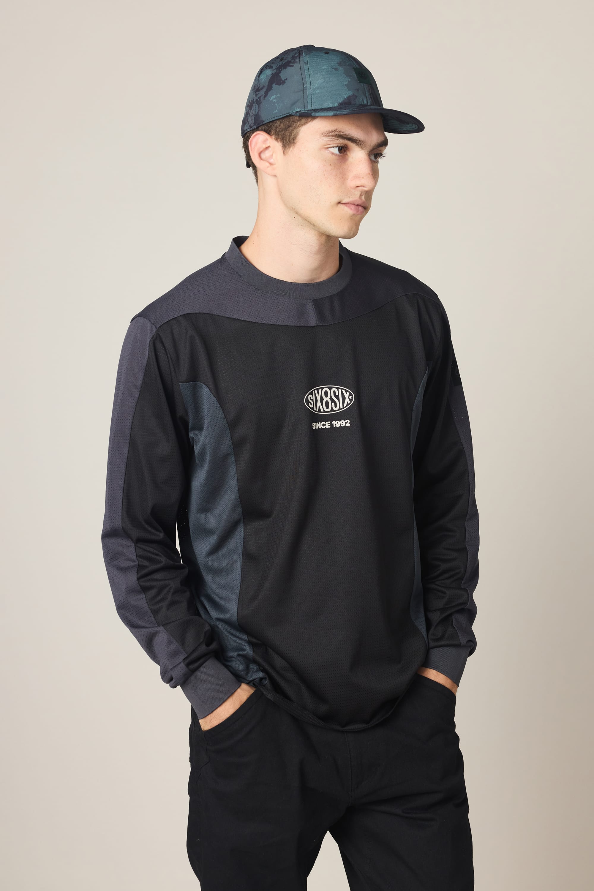 A young man in a blue camo cap, black pants, and the 686 Men's 92 Turbo LS Tech Tee by 686 stands with hands in pockets against a neutral background.