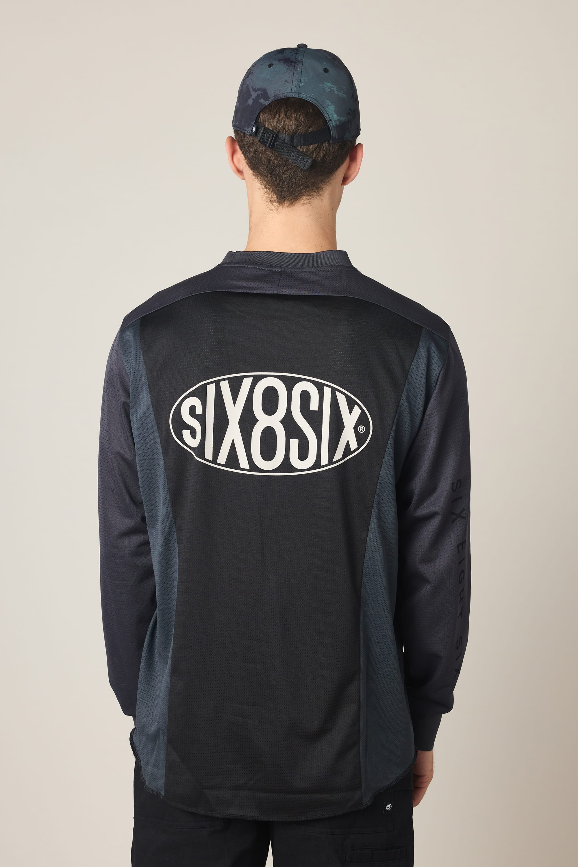A person is photographed from behind against a plain background, wearing the 686 Men's 92 Turbo LS Tech Tee featuring SIX8SIX in a large white oval logo on the back, plus a blue patterned baseball cap.