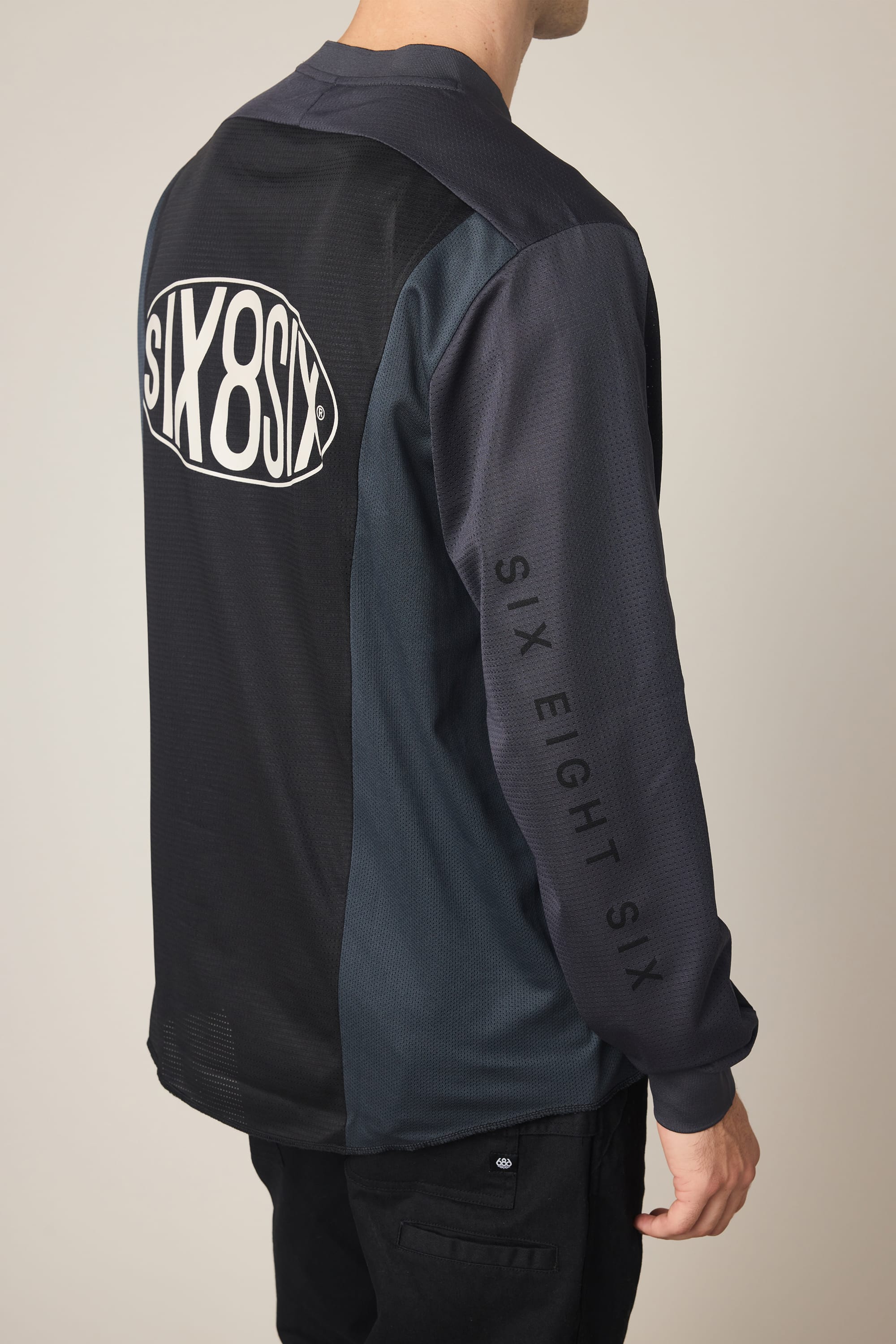 A person wearing a 686 Men's 92 Turbo LS Tech Tee, black polyester with "SIX8SIX" in a white oval on the back and "SIX EIGHT SIX" printed vertically on the sleeve, stands against a plain light background.