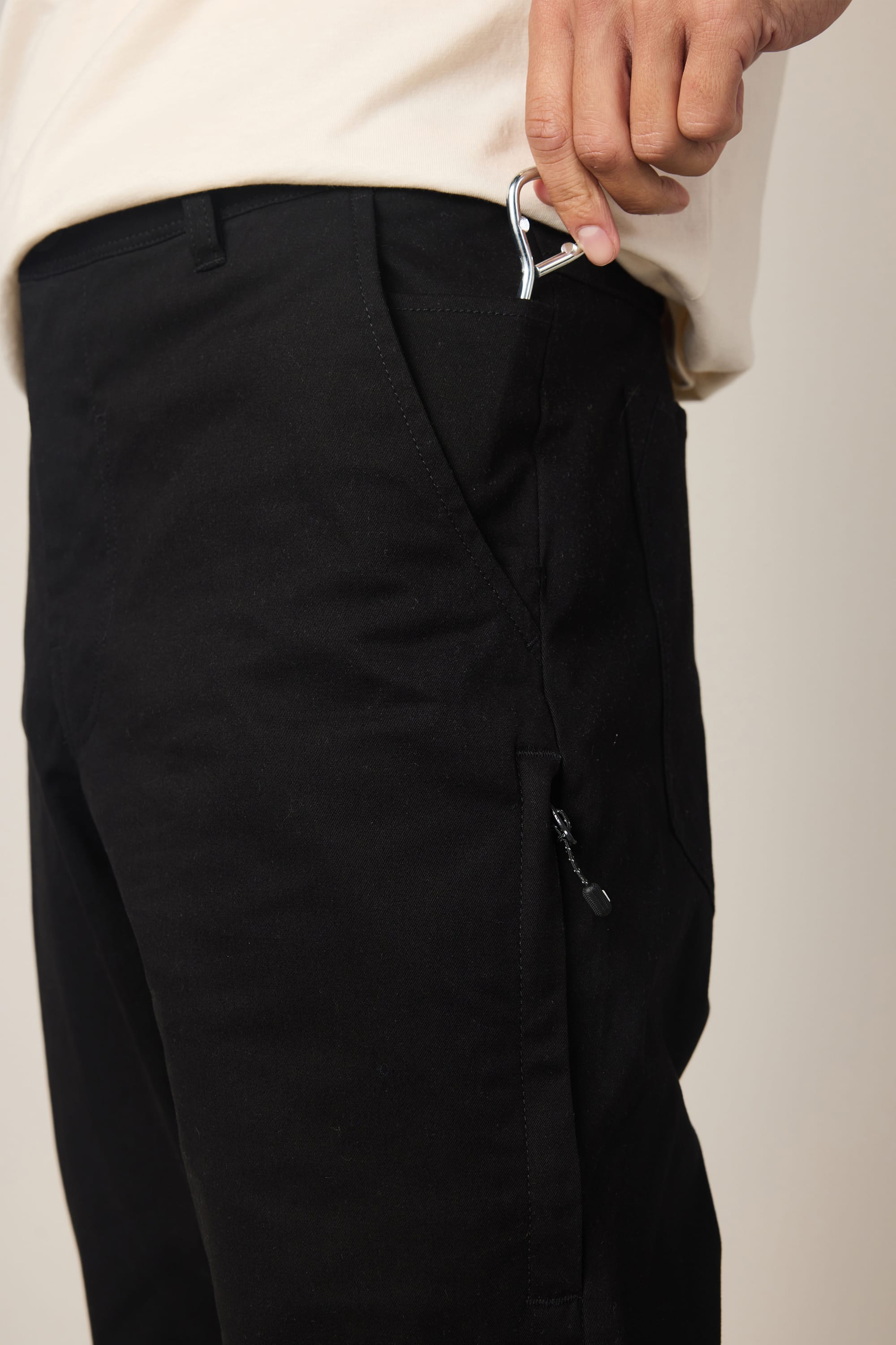 Wearing the 686 Men's Everywhere® Standard Pant - Slim Fit in black, a person places a small metal carabiner into their front pocket while sporting a beige shirt.