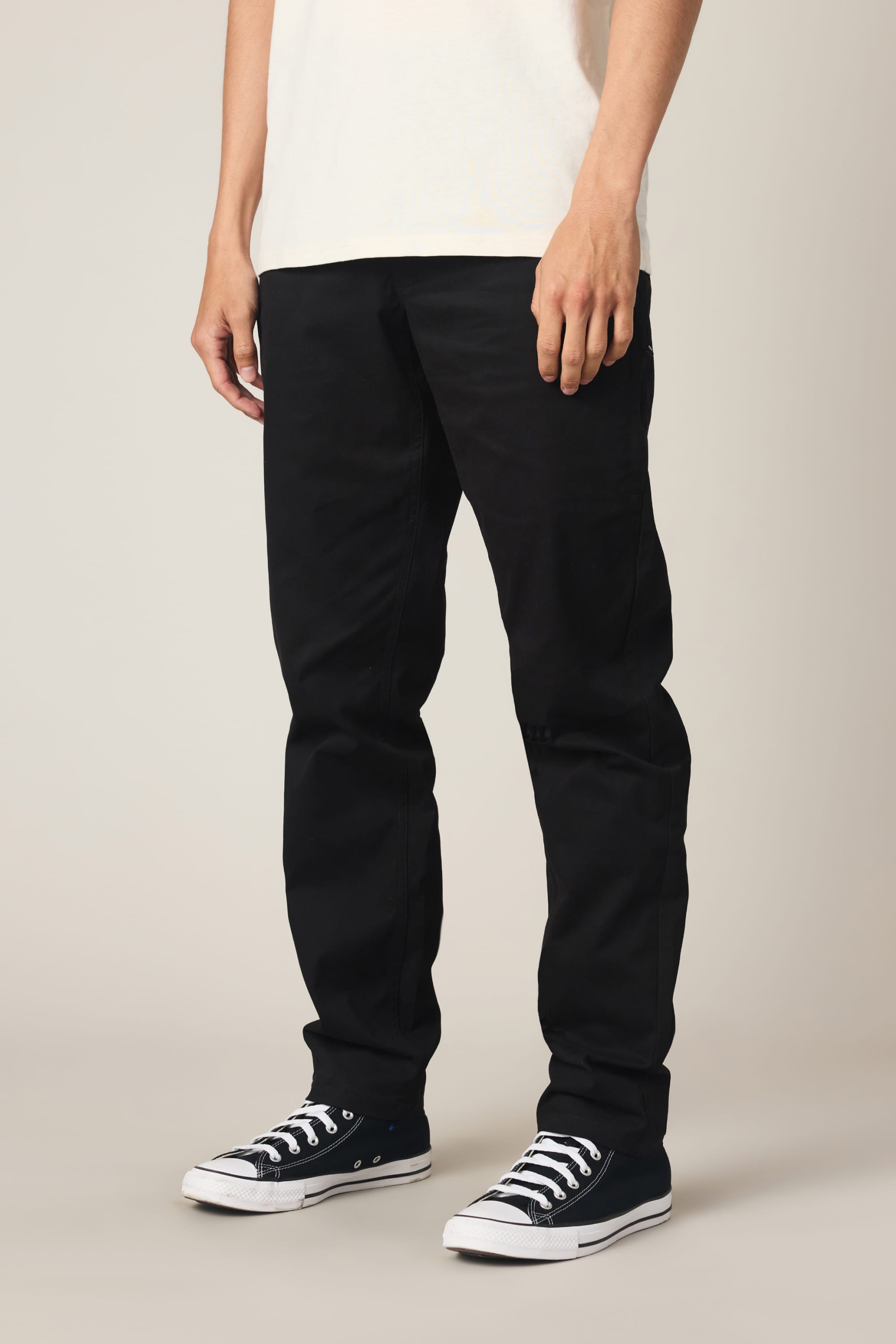 A person wearing a white shirt, 686 Men's Everywhere® Standard Pant - Slim Fit in black by 686, and black low-top sneakers stands against a plain background, visible from shoulders down.