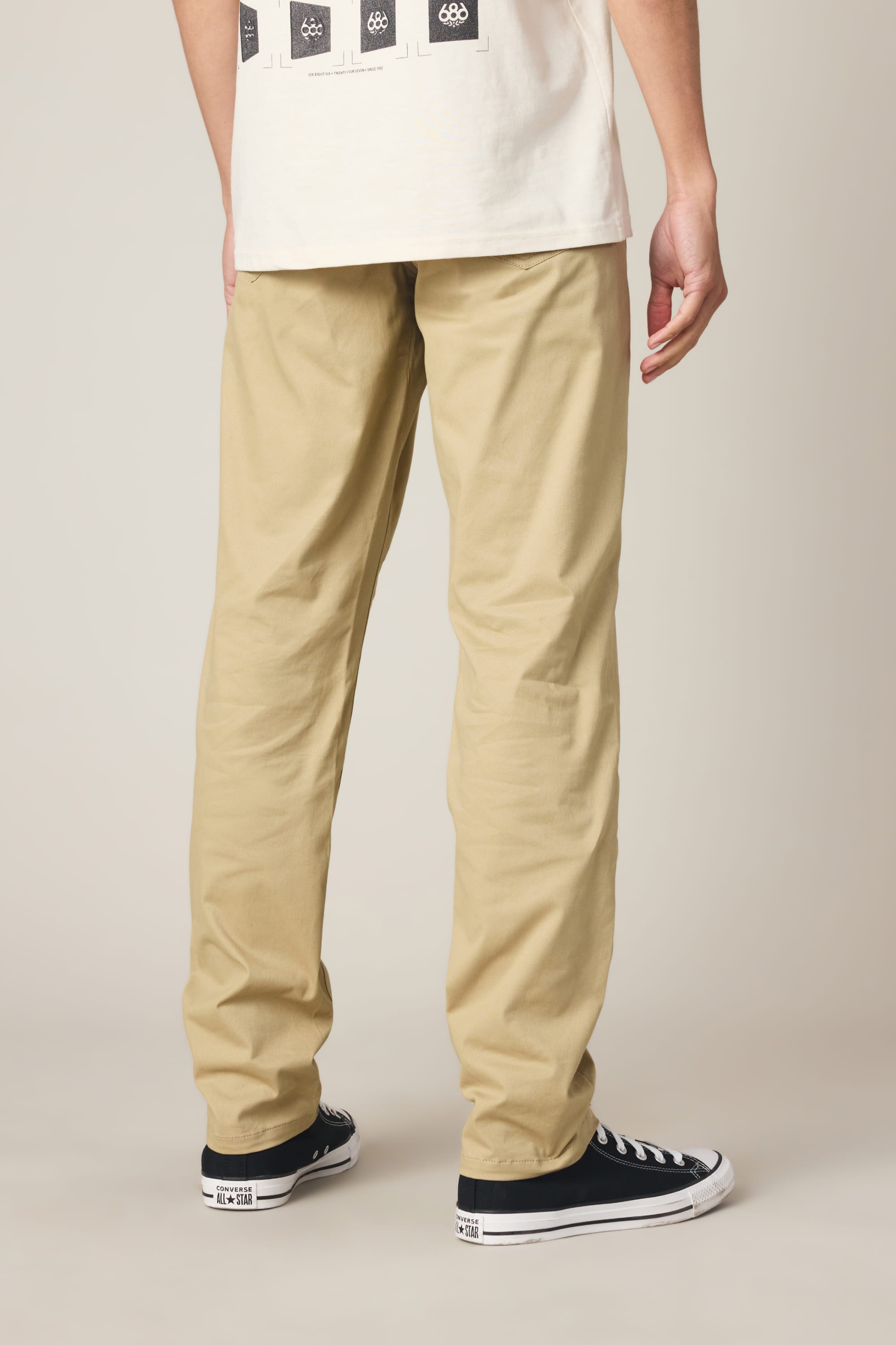 A person stands with their back to the camera, wearing the 686 Men's Everywhere® Standard Pant - Slim Fit in khaki. Only their lower body is shown against a neutral background, paired with a white shirt and black low-top sneakers with white soles.