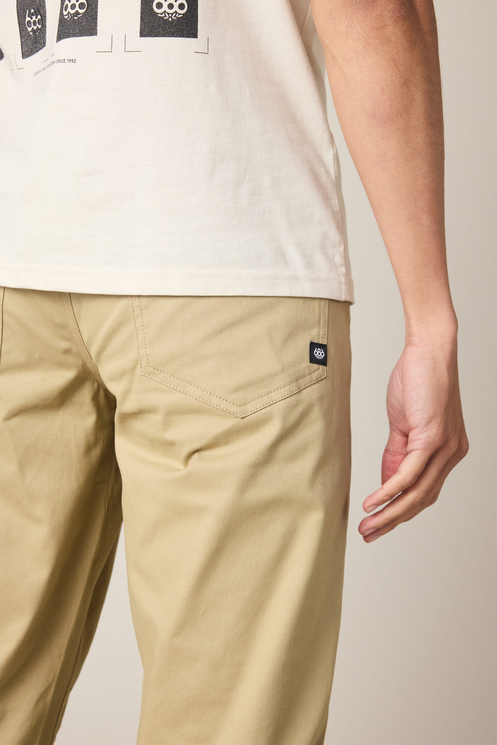 A person is shown from the back and side wearing a white T-shirt and 686 Men's Everywhere® Standard Pant - Slim Fit in beige, with their right arm relaxed by their side and a small black tag visible on the back pocket.