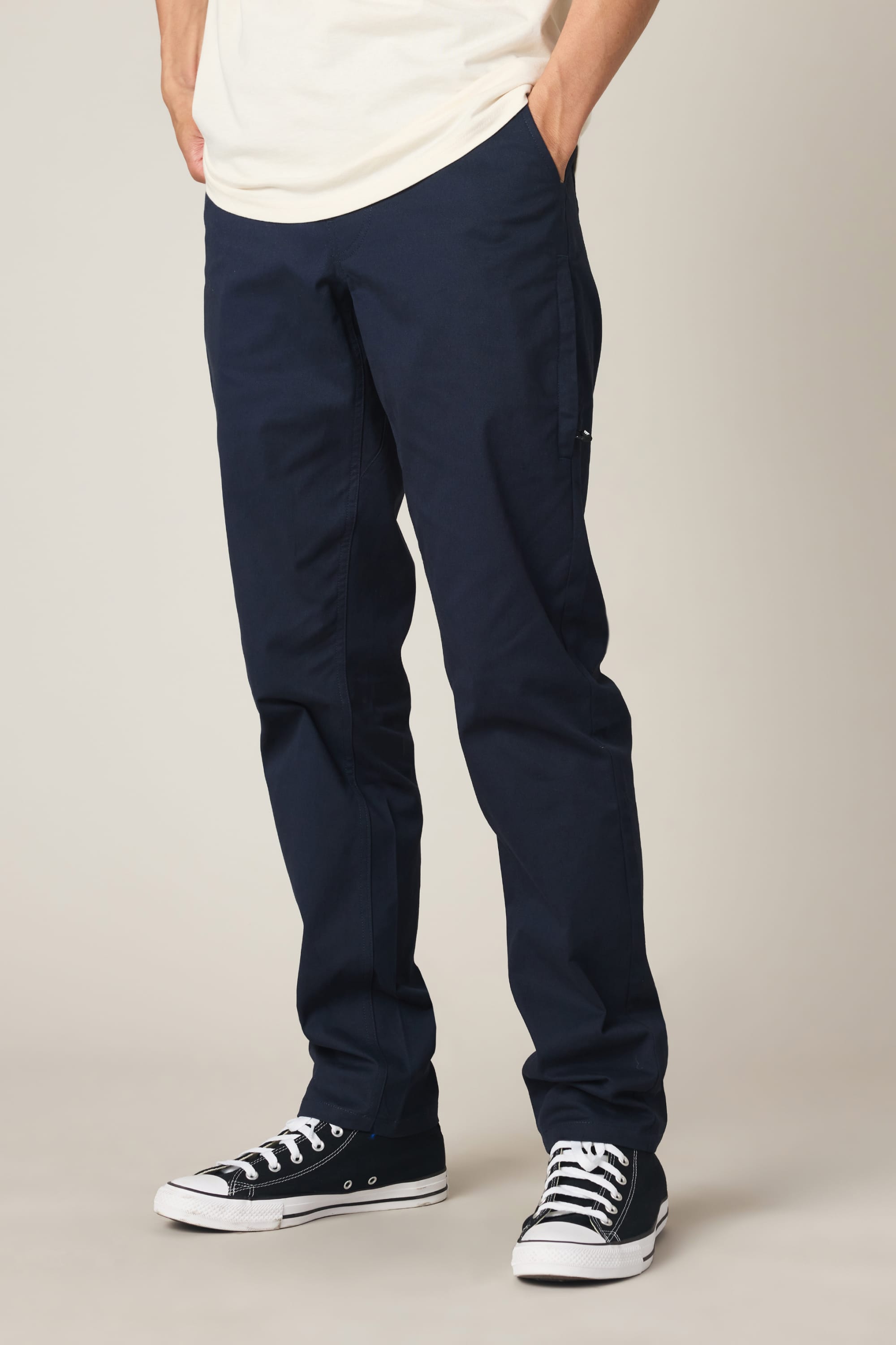 A person wearing 686 Men's Everywhere® Standard Pant - Slim Fit in navy blue, a white shirt, and black low-top sneakers with white soles stands with hands in pockets against a plain light background.