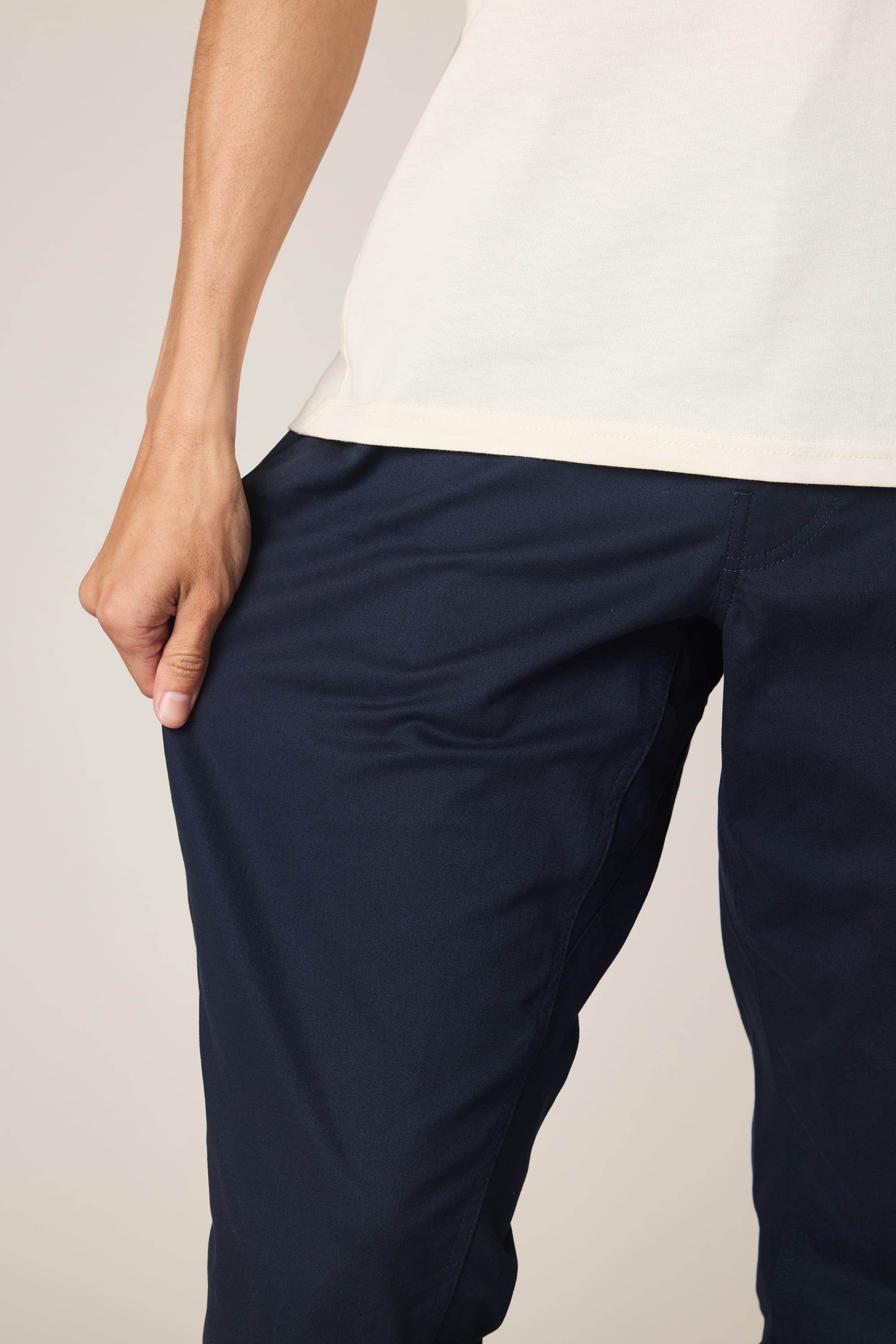 A person wears a white shirt and 686 Men's Everywhere® Standard Pant - Slim Fit in dark blue, holding the pant to highlight its texture and fit. Only the torso and legs are visible in the image.