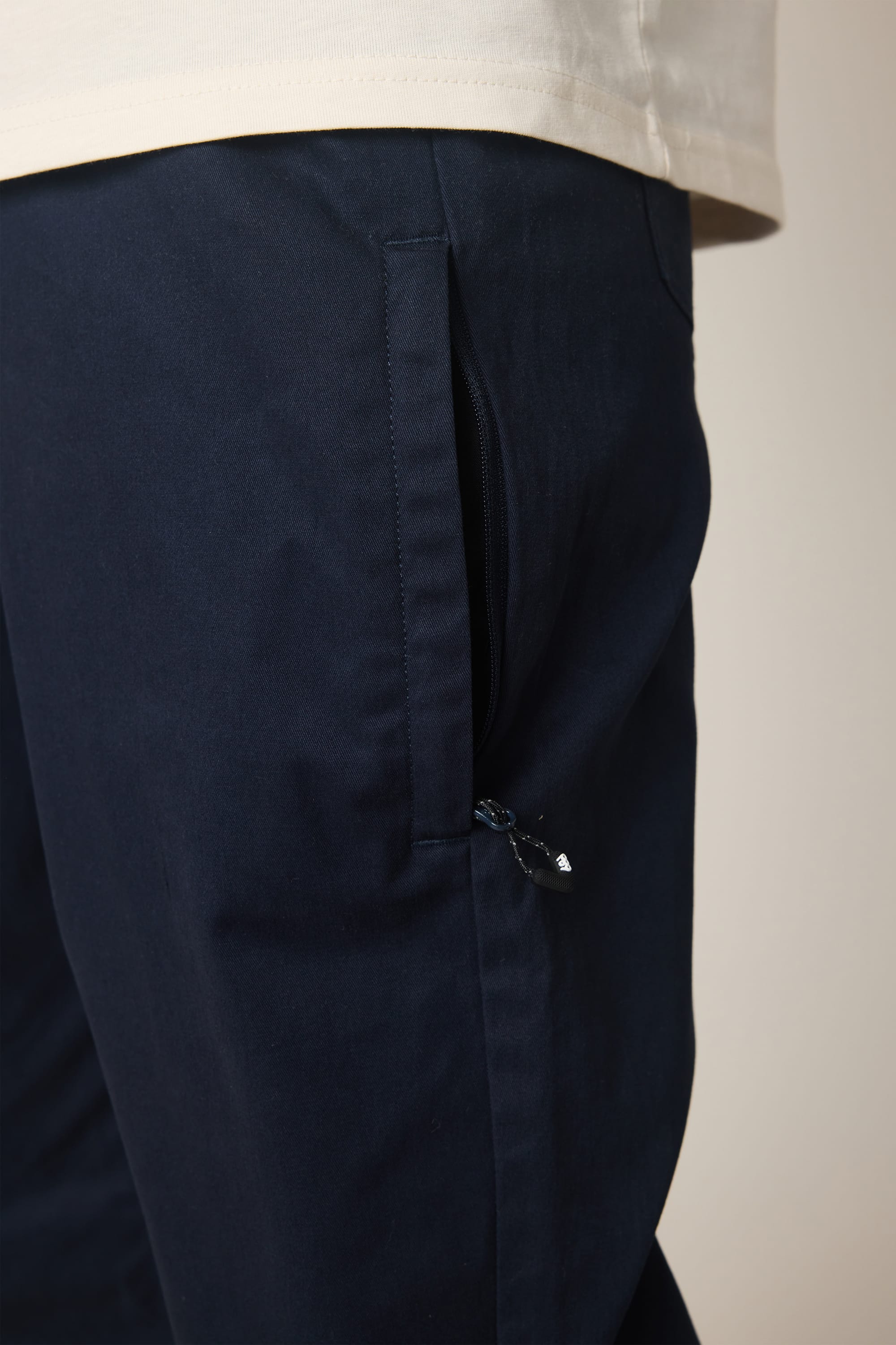 A close-up of a person wearing the 686 Men's Everywhere® Standard Pant - Relaxed Fit in navy blue, featuring a zippered side pocket, paired with a light beige shirt.