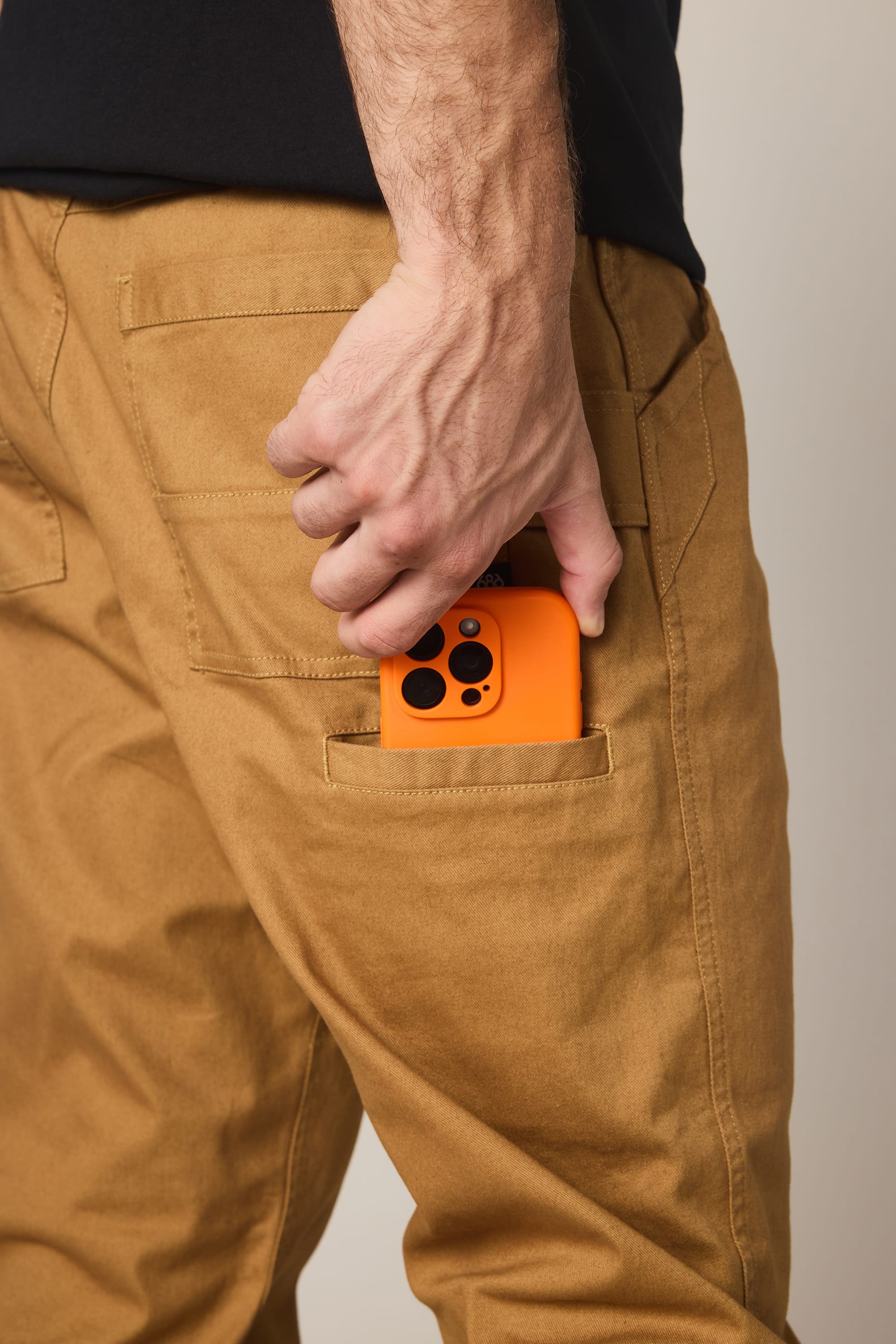 A person wearing 686 Men's Unwork® Tradesman Pant - Relaxed Fit places an orange smartphone with a black camera case into the back pocket.